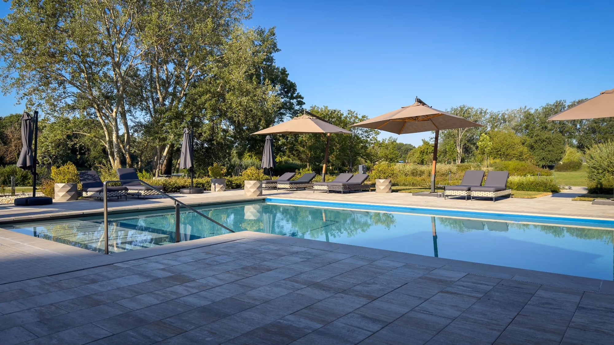 Outdoor pool with sun loungers, red flowers, green lawn, trees, and forested mountains in background.
