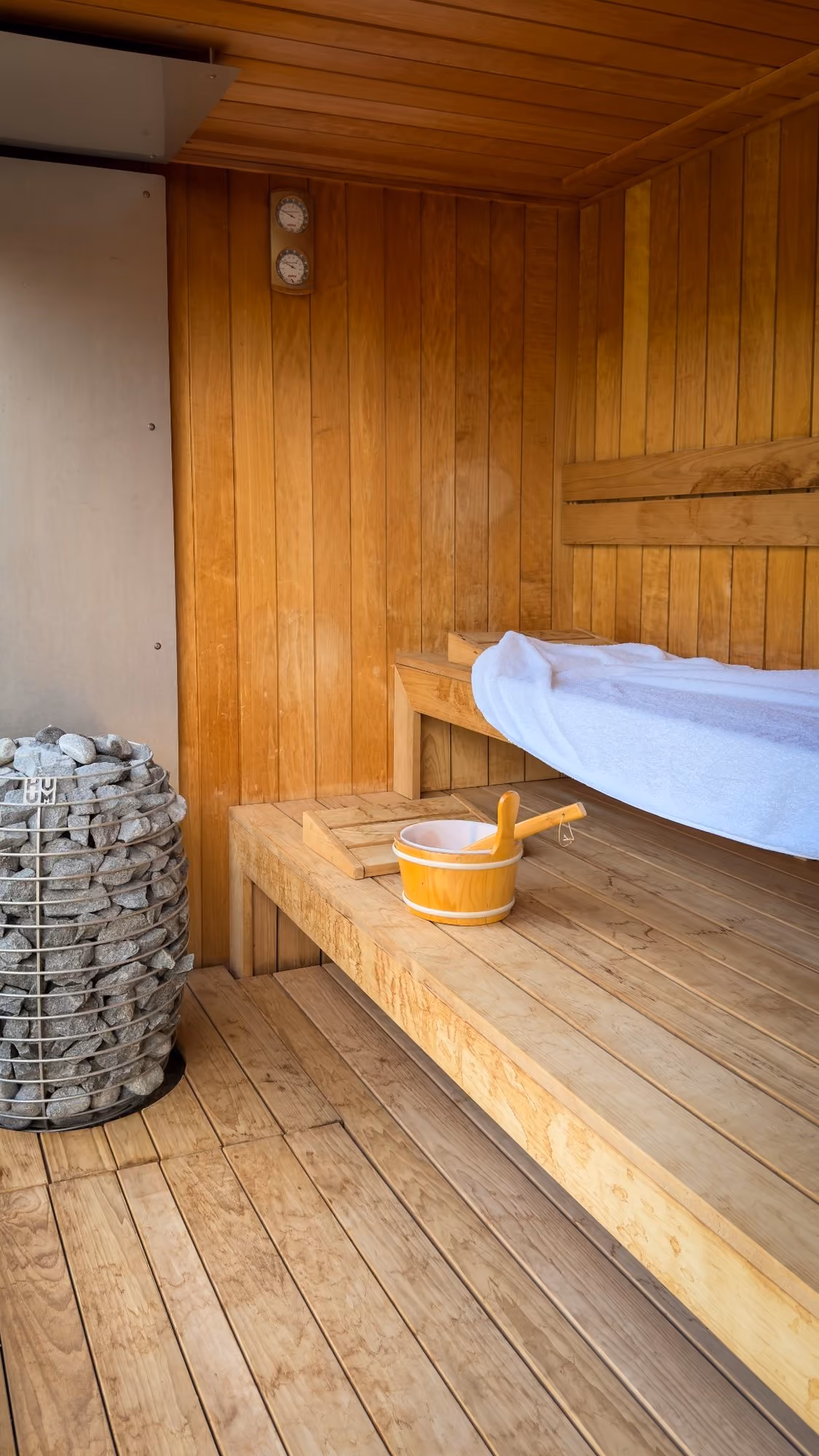Wood-paneled sauna interior with bench, towel, wooden bucket, ladle, and heater with stones.