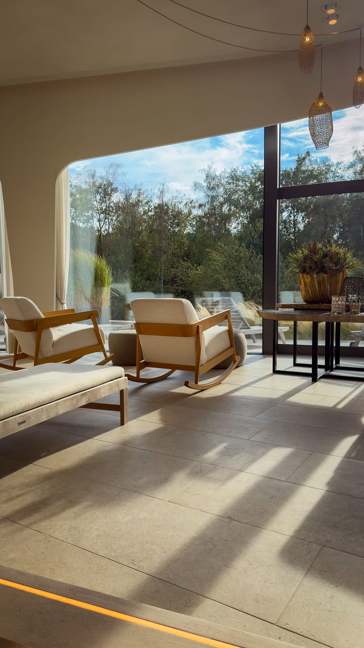 Modern living room with beige cushioned wooden rocking chairs and loungers near large floor-to-ceiling windows showing green trees outside, illuminated by natural sunlight casting shadows on tiled floor.