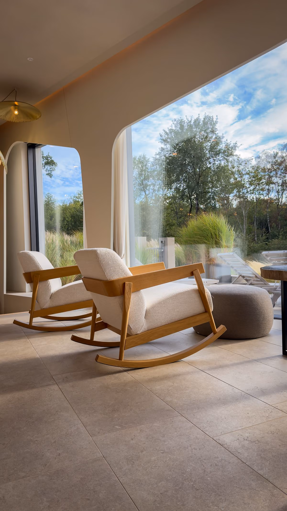 Modern lounge area with natural wood round coffee tables, light gray sofas, and large glass windows revealing greenery outside.