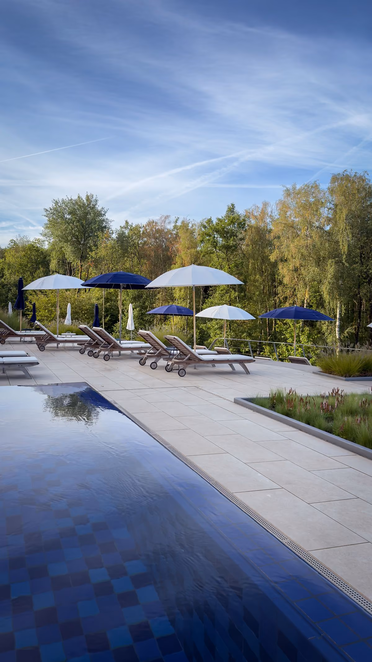 Outdoor pool beside a tiled deck with multiple lounge chairs under alternating white and dark blue umbrellas, surrounded by green trees under a clear blue sky.