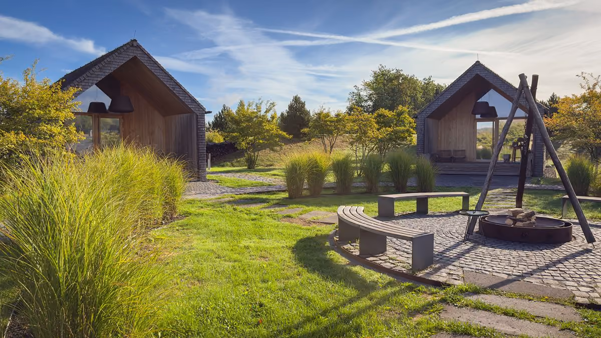 A serene outdoor setting with two modern wooden cabins, green grass, shrubs, and a circular stone fire pit area with a tripod over it and curved benches around.