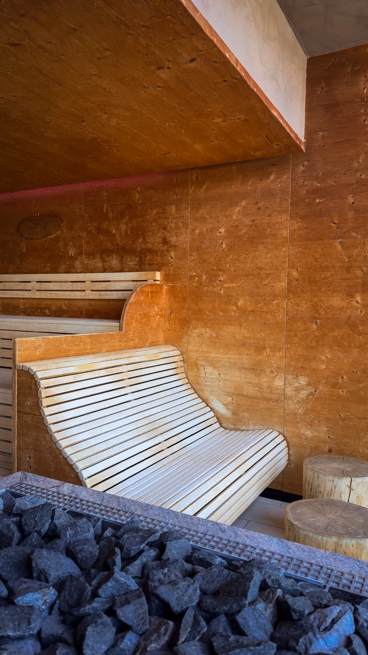 Wood-paneled sauna interior with a curved wooden bench, wooden stools, and black sauna stones in the foreground.