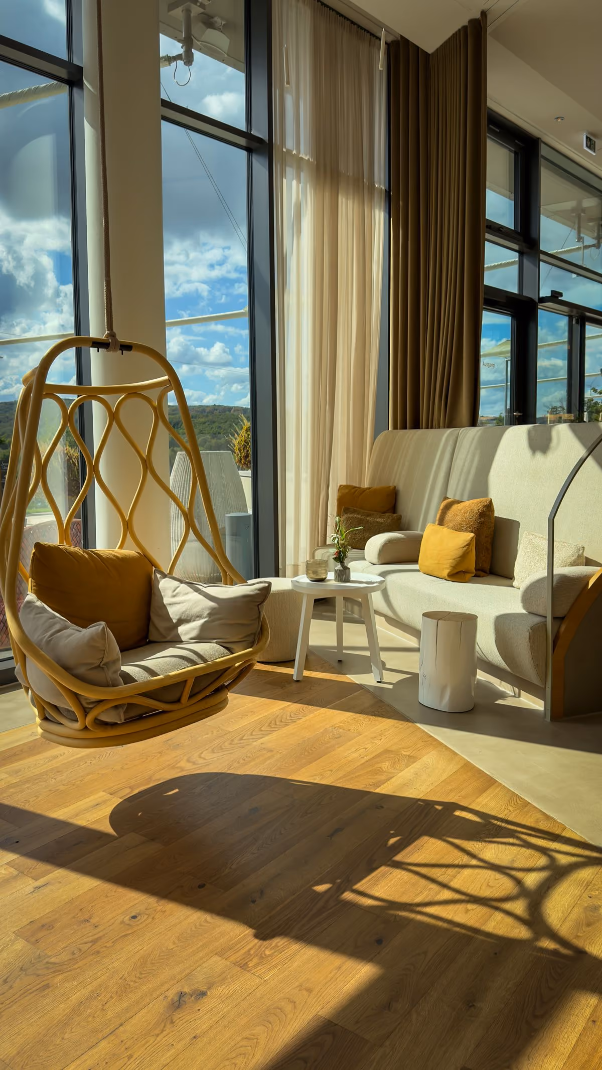 Sunlit modern living room with large windows, featuring a hanging wicker chair with cushions, a beige sofa with mustard and beige pillows, a small round white side table with a plant, and a wooden floor casting shadows.