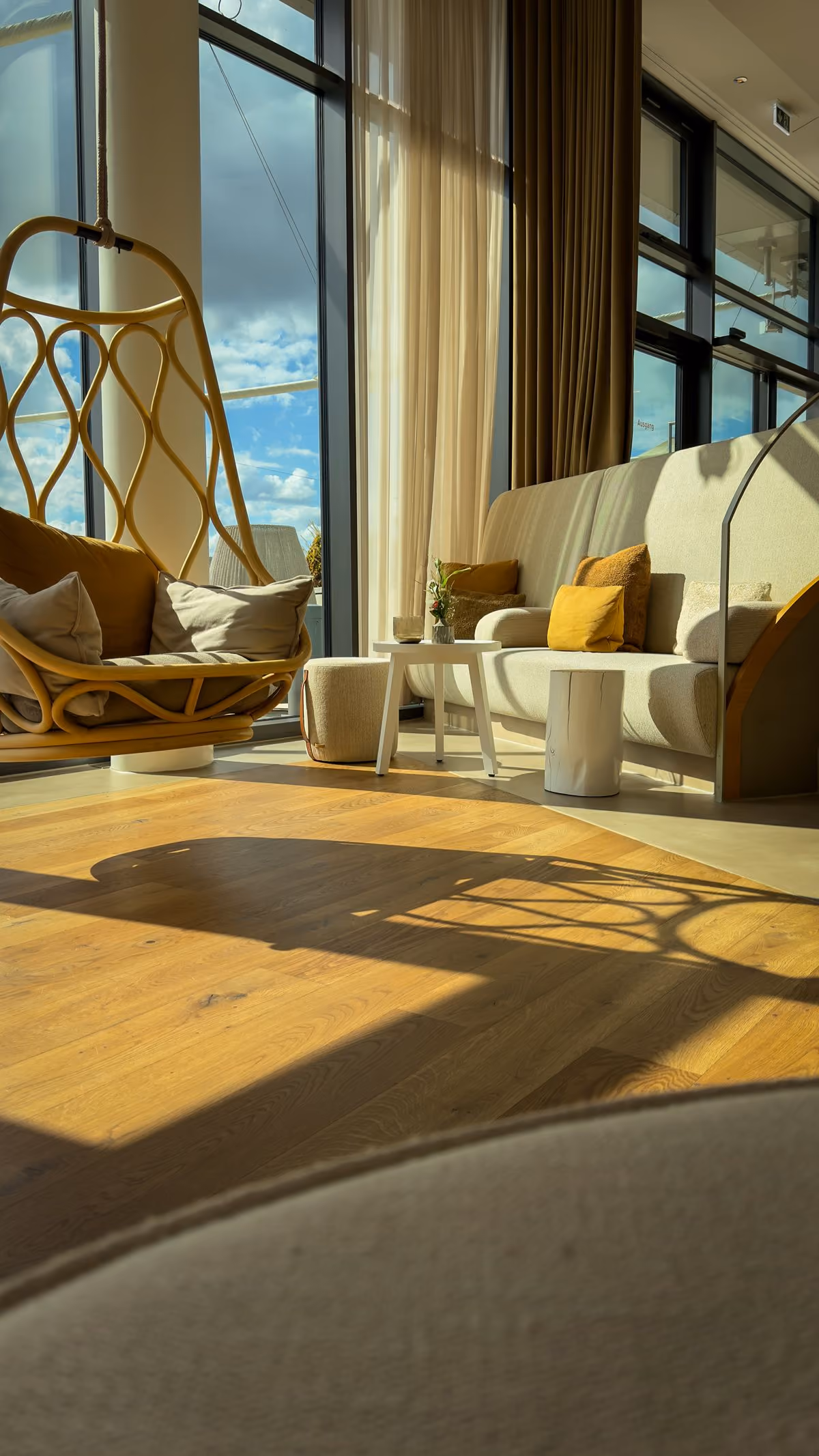 Sunlit modern living space with large floor-to-ceiling windows, a hanging wicker chair with cushions, a sofa with yellow and beige pillows, light wooden floors, beige curtains, and small white tables and stools.