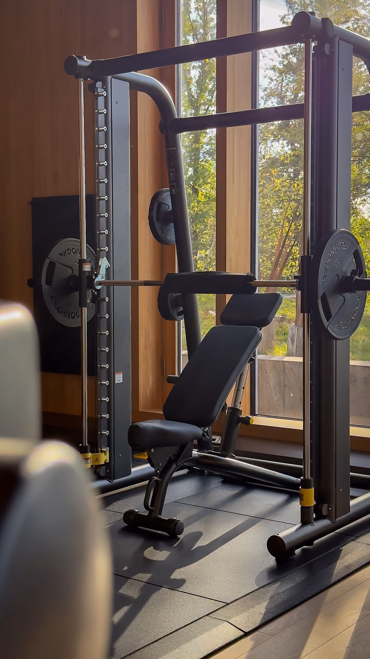Modern Pilates reformer machine with wood frame and padded platform in a bright studio with wooden floor and dark textured wall.