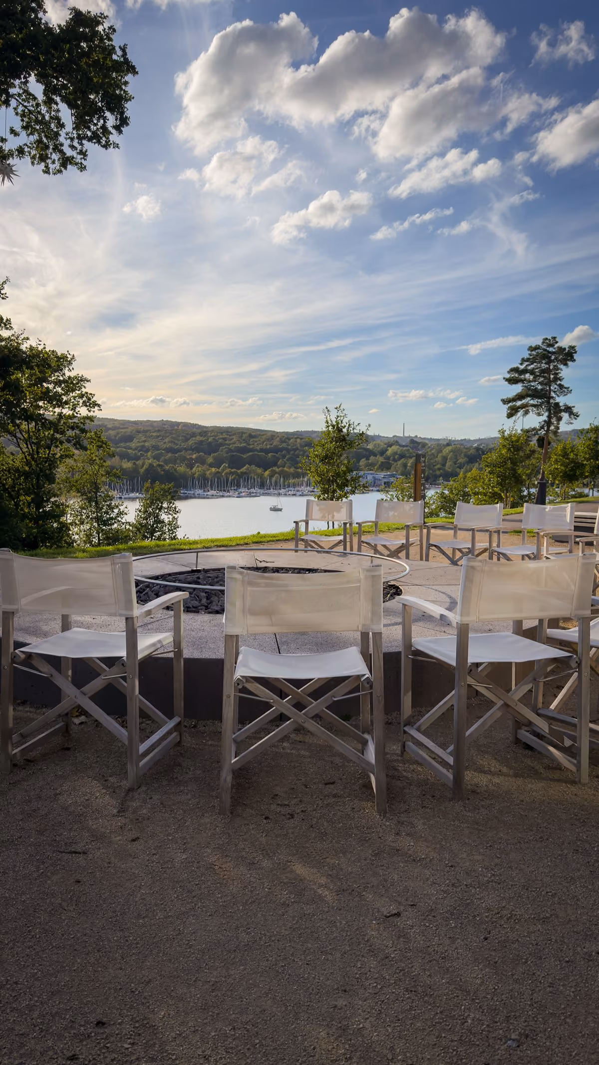 Outdoor seating area with white director-style chairs arranged around a circular fire pit, overlooking a scenic view of a lake with boats and forested hills under a partly cloudy sky.