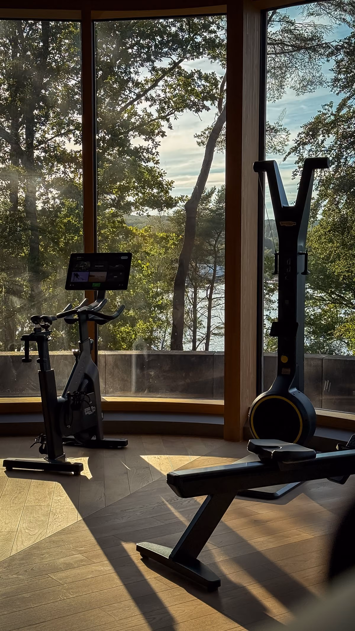 Indoor exercise room with stationary bike, rowing machine, and workout bench in front of large curved windows overlooking trees and a body of water outside in natural daylight.