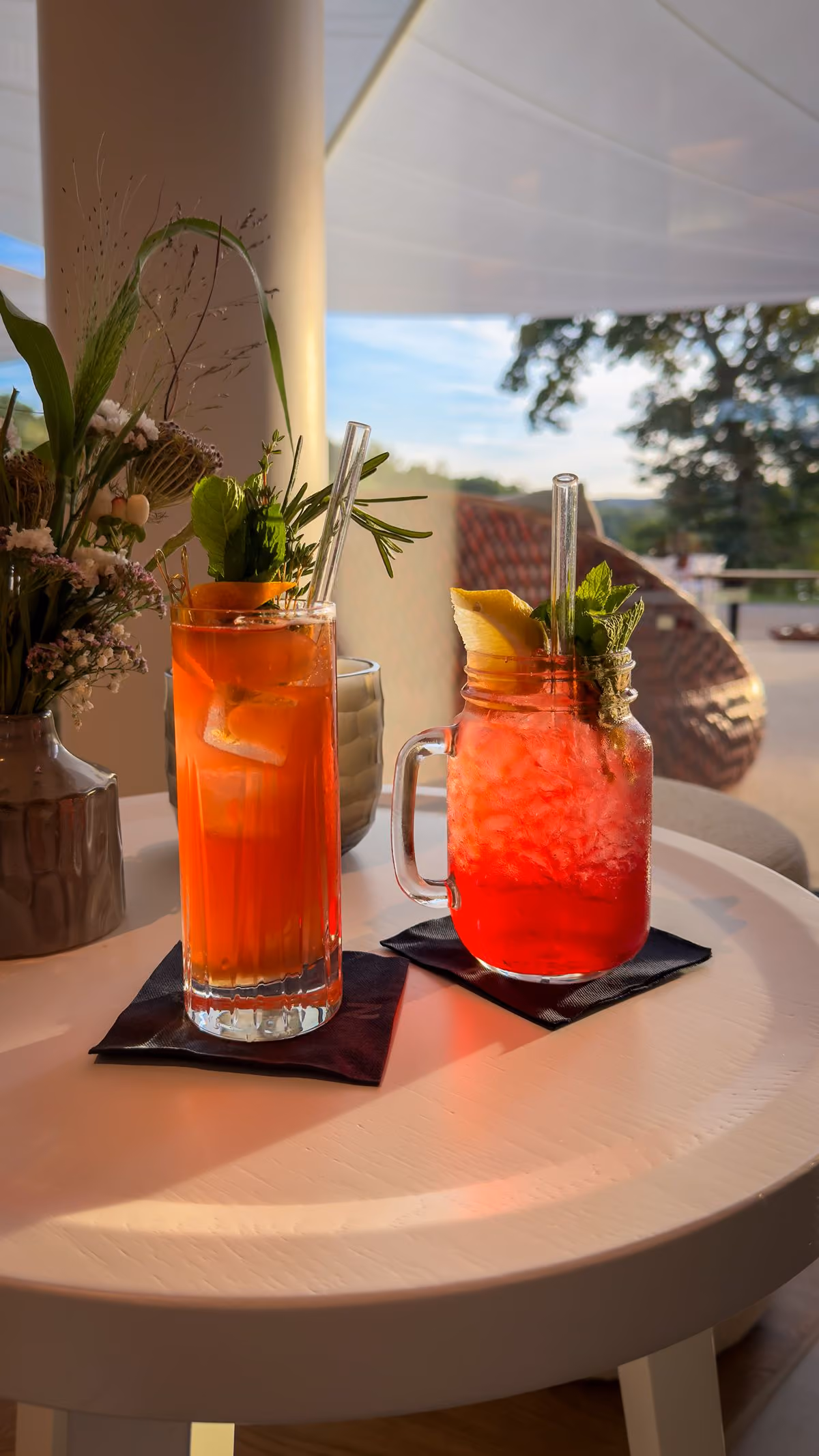Two refreshing cocktails on a round white table; the left glass is tall with orange liquid, ice, herbs, and a straw, the right is a mason jar with red crushed ice, lemon slice, mint, and a straw, set against an outdoor background with wicker furniture and greenery.