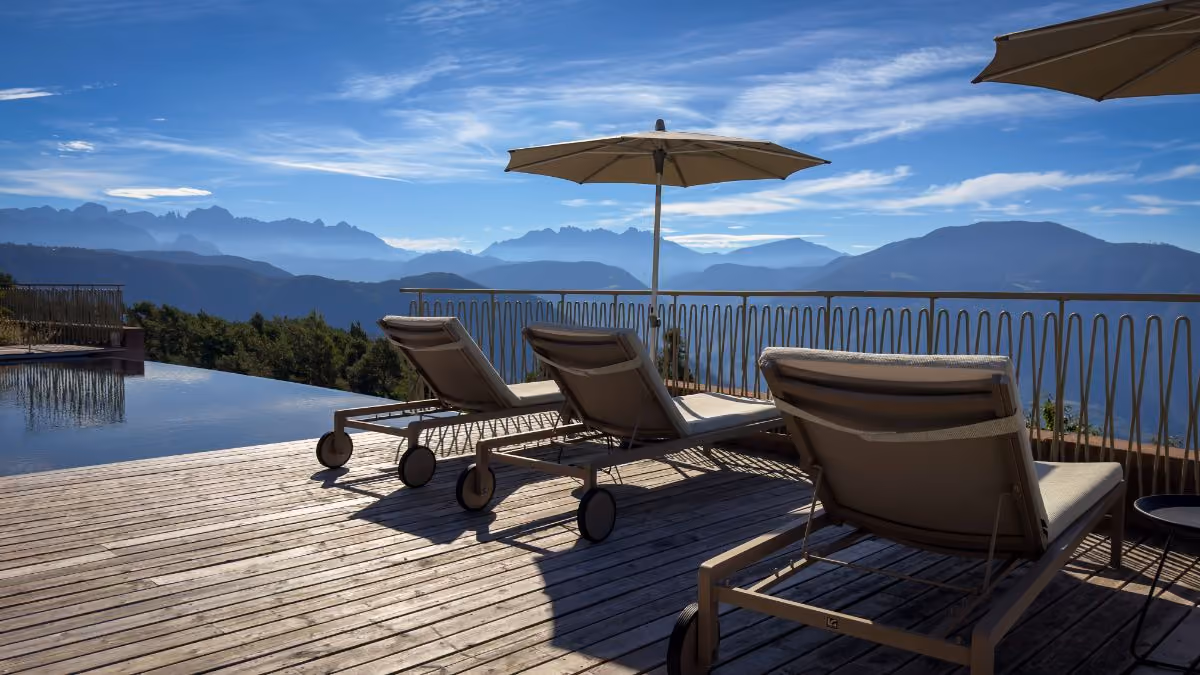 Three lounge chairs on a wooden deck beside a pool with umbrellas and a mountain range under a partly cloudy blue sky in the background.