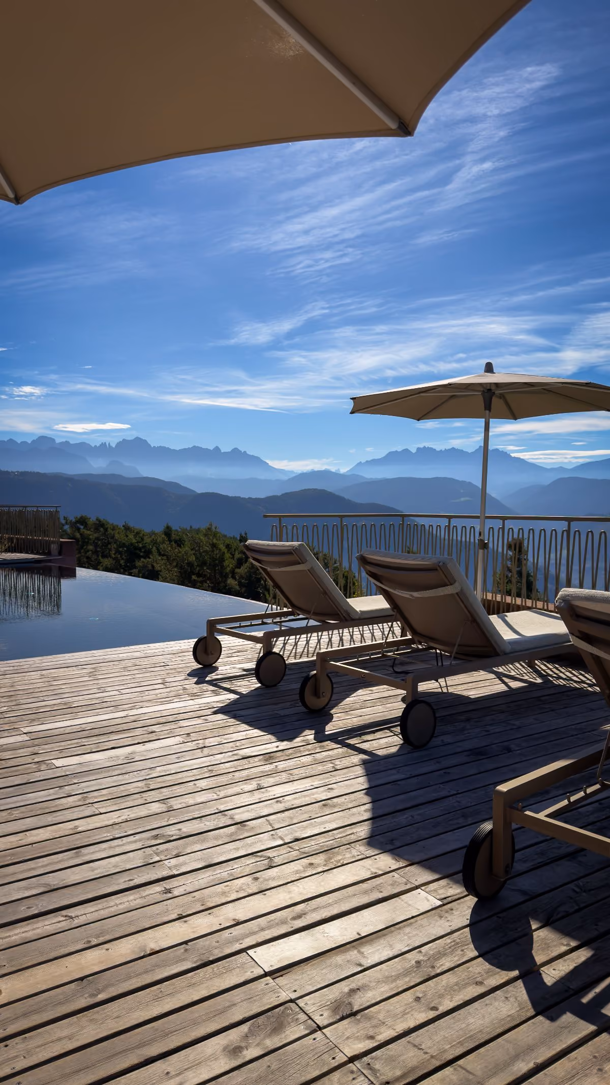 Wooden deck with sun loungers and umbrellas beside an infinity pool overlooking distant mountain ranges under a blue sky.