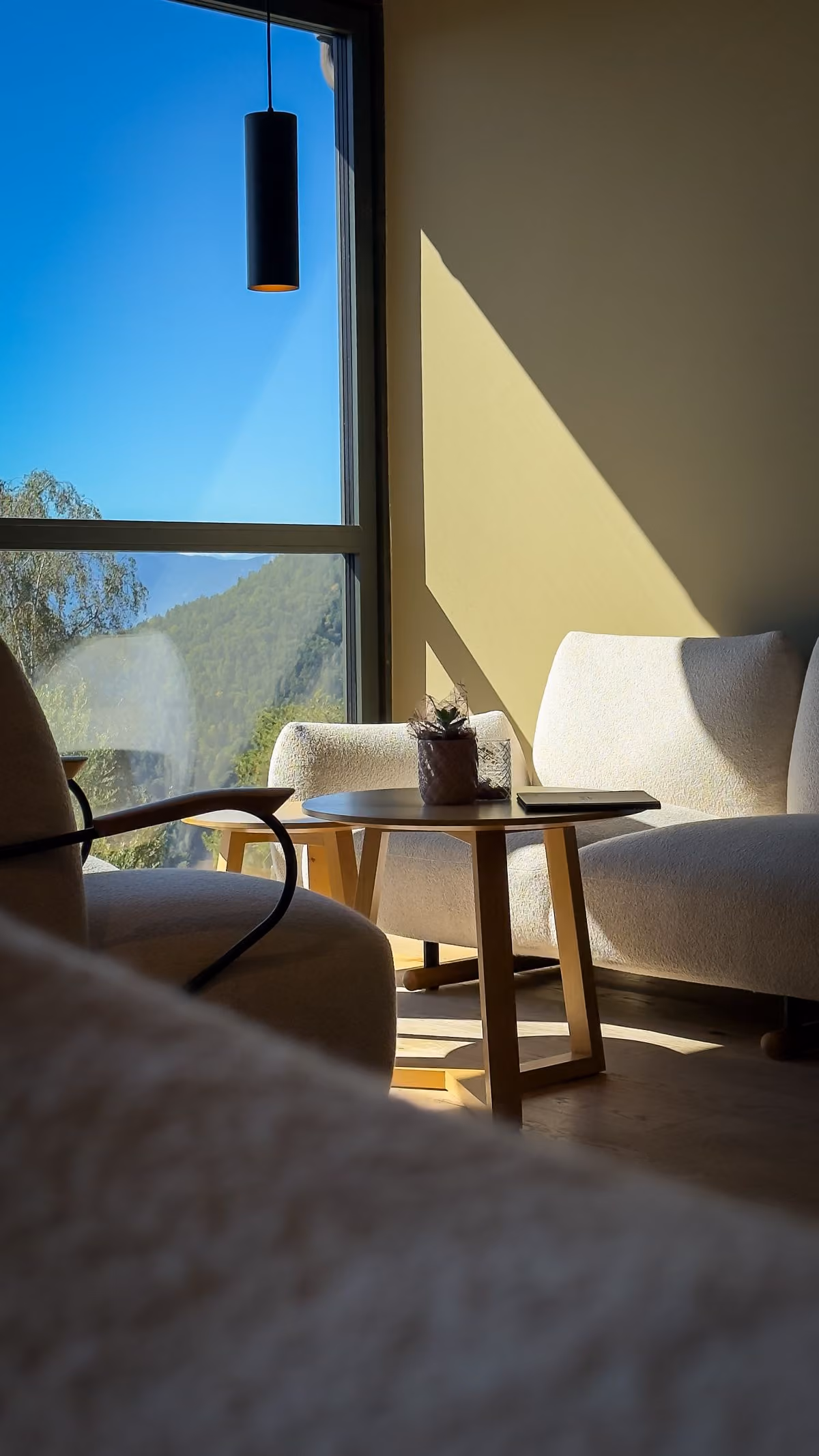 Bright cozy corner with modern white armchairs around a round wooden table holding a small potted succulent and a closed laptop, next to a large window showing clear blue sky and greenery outside.