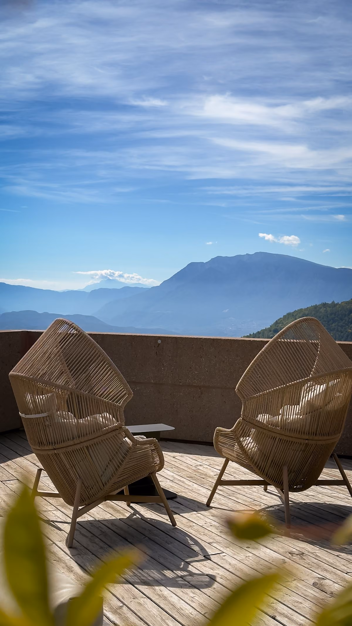 Two wicker lounge chairs on a wooden deck facing a concrete railing with a scenic view of mountains under a blue sky with wispy clouds.