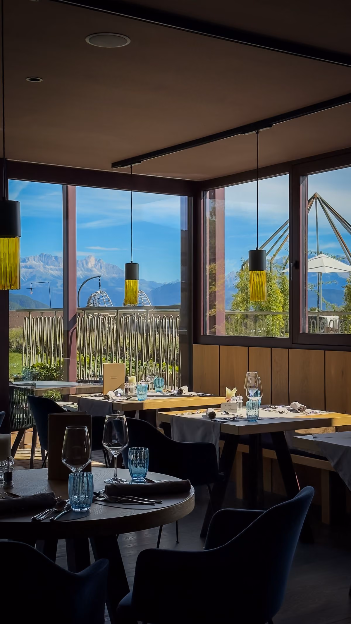 A modern restaurant interior with neatly set dining tables featuring glassware, napkins, and cutlery, large windows offering a view of mountains and outdoor seating with umbrellas under a clear sky.