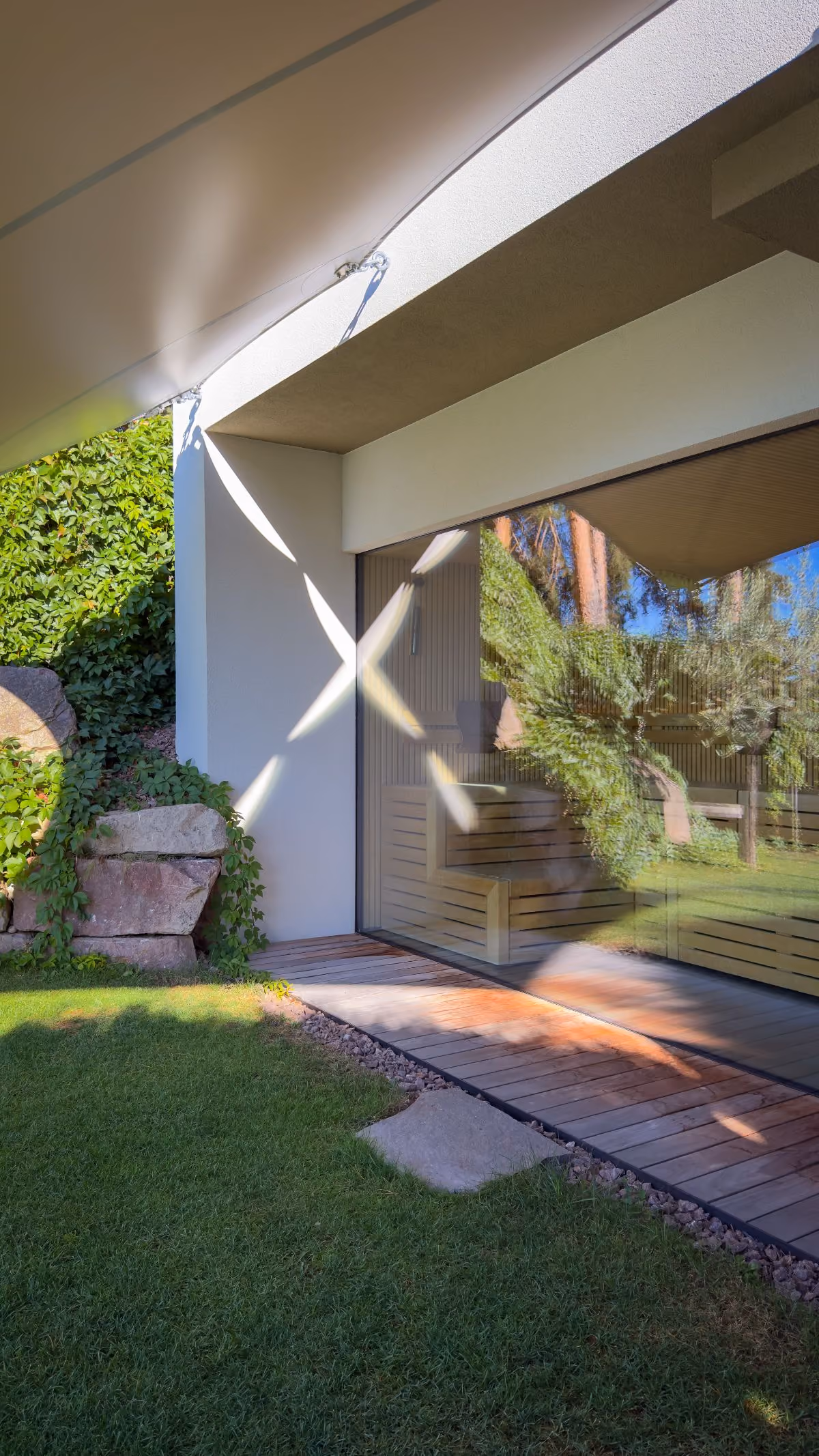 Modern outdoor patio area with wooden decking next to green grass, stone steps covered in ivy, and a large glass window reflecting trees and sunlight.
