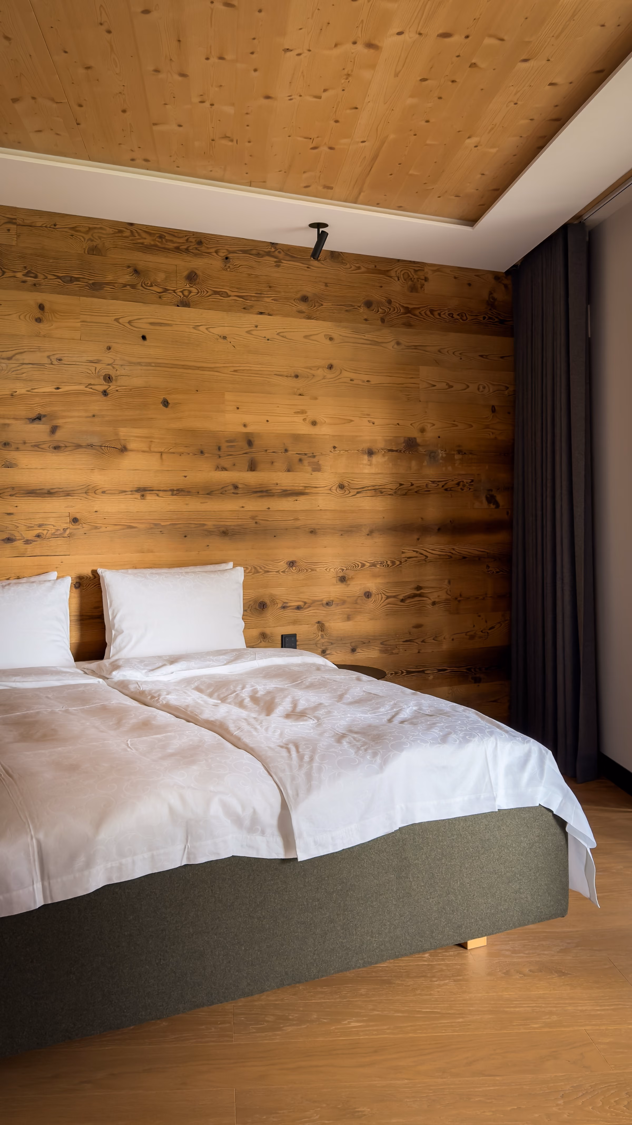 A neatly made bed with white bedding and pillows set against a wooden paneled wall, next to dark curtains and a wooden floor.