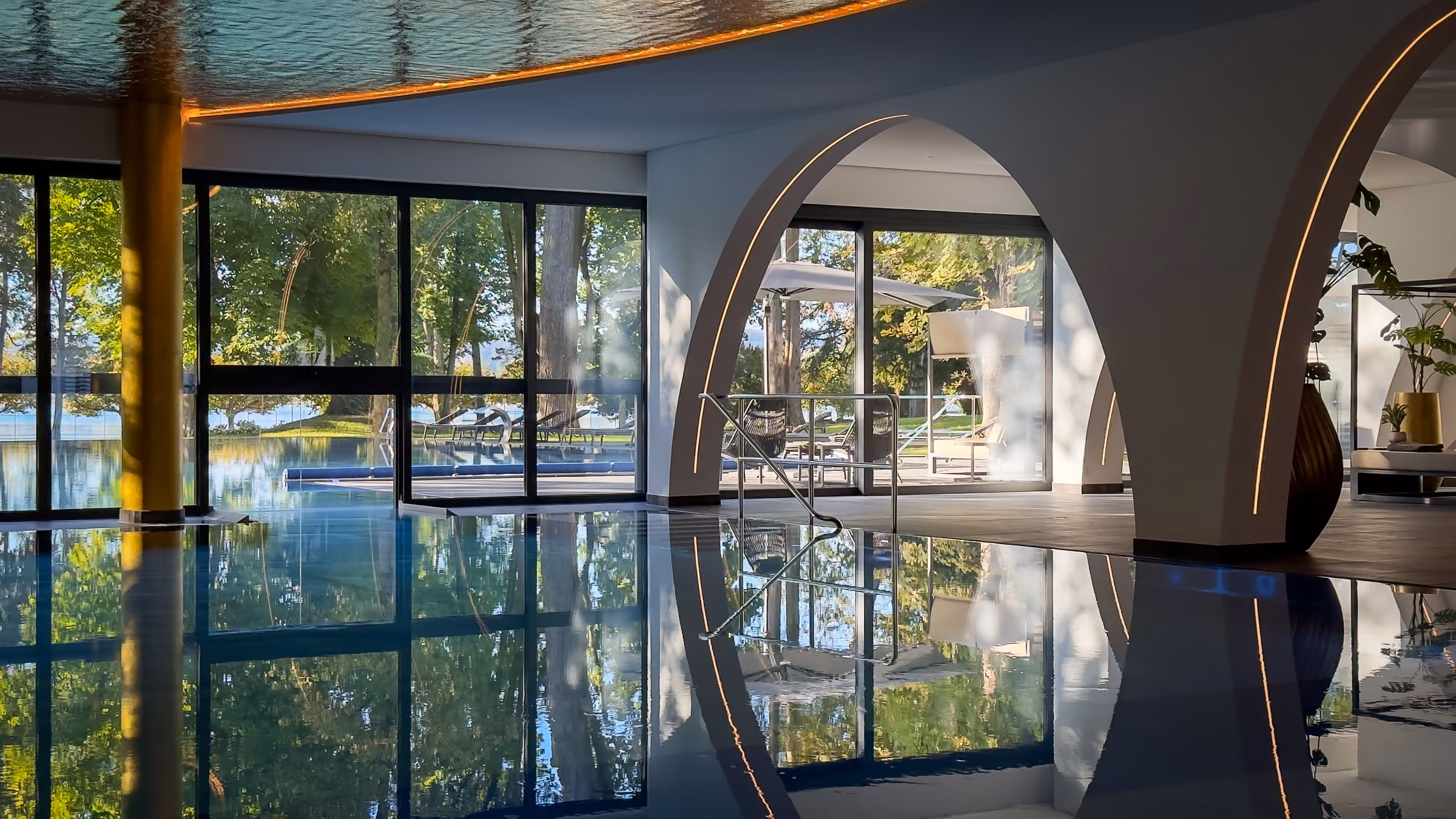 Indoor pool area with large arched windows reflecting the surrounding trees and outdoor patio furniture, creating a serene atmosphere.