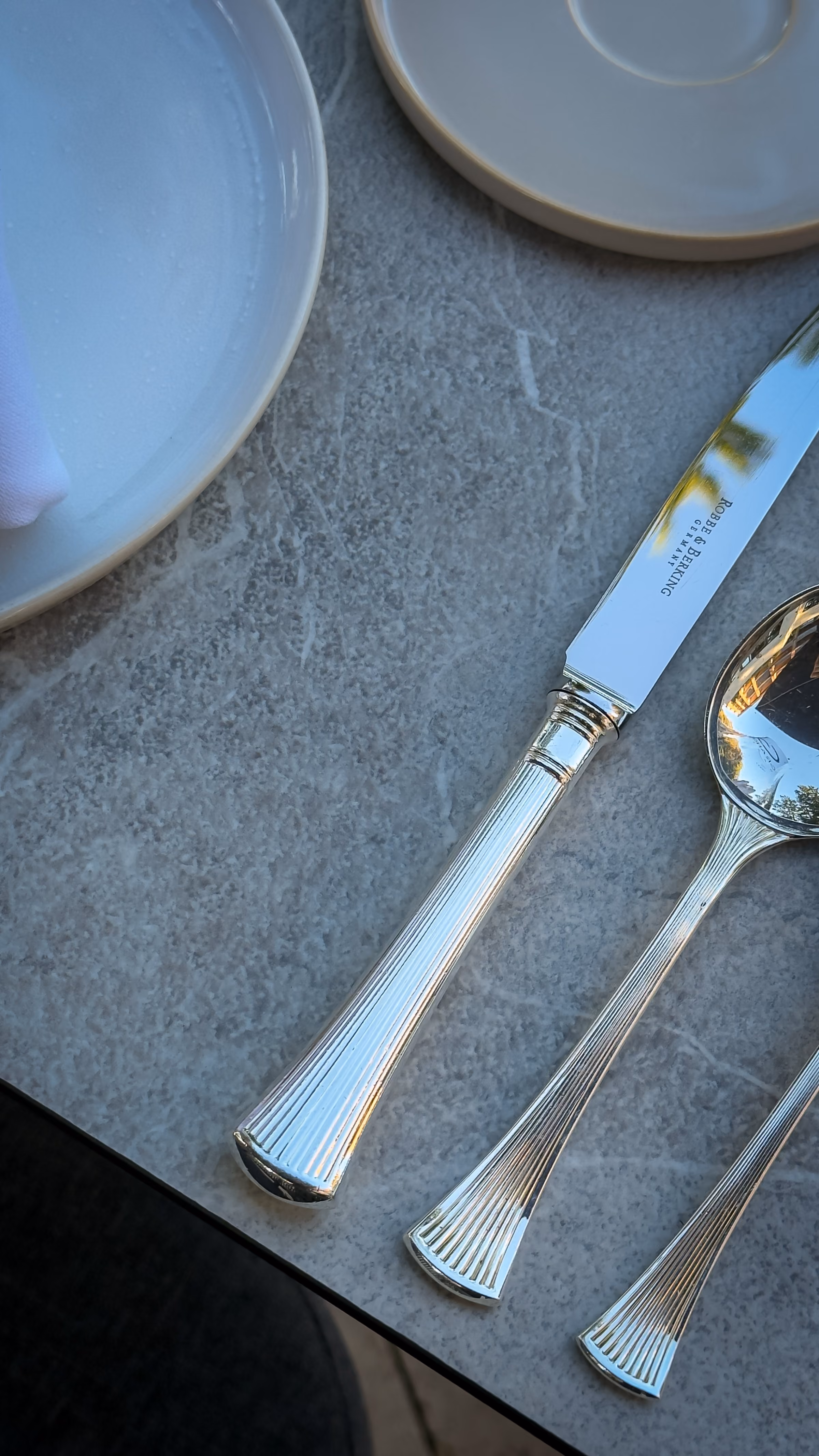 Close-up of a polished silver knife and spoon with detailed handles on a gray marble table next to a beige plate and saucer.