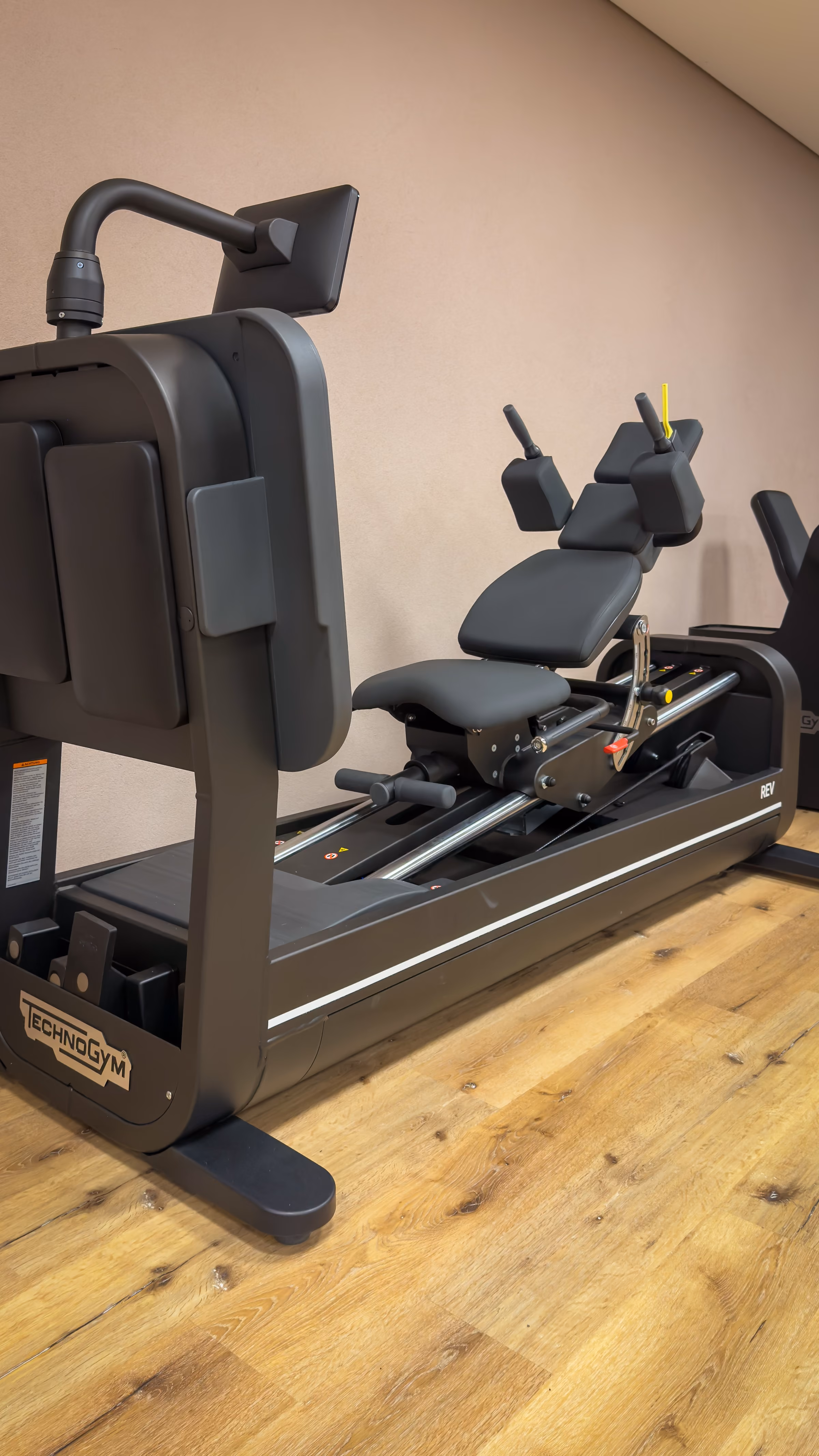 Technogym rowing machine with black padded seat and adjustable handles on wooden floor against beige wall.