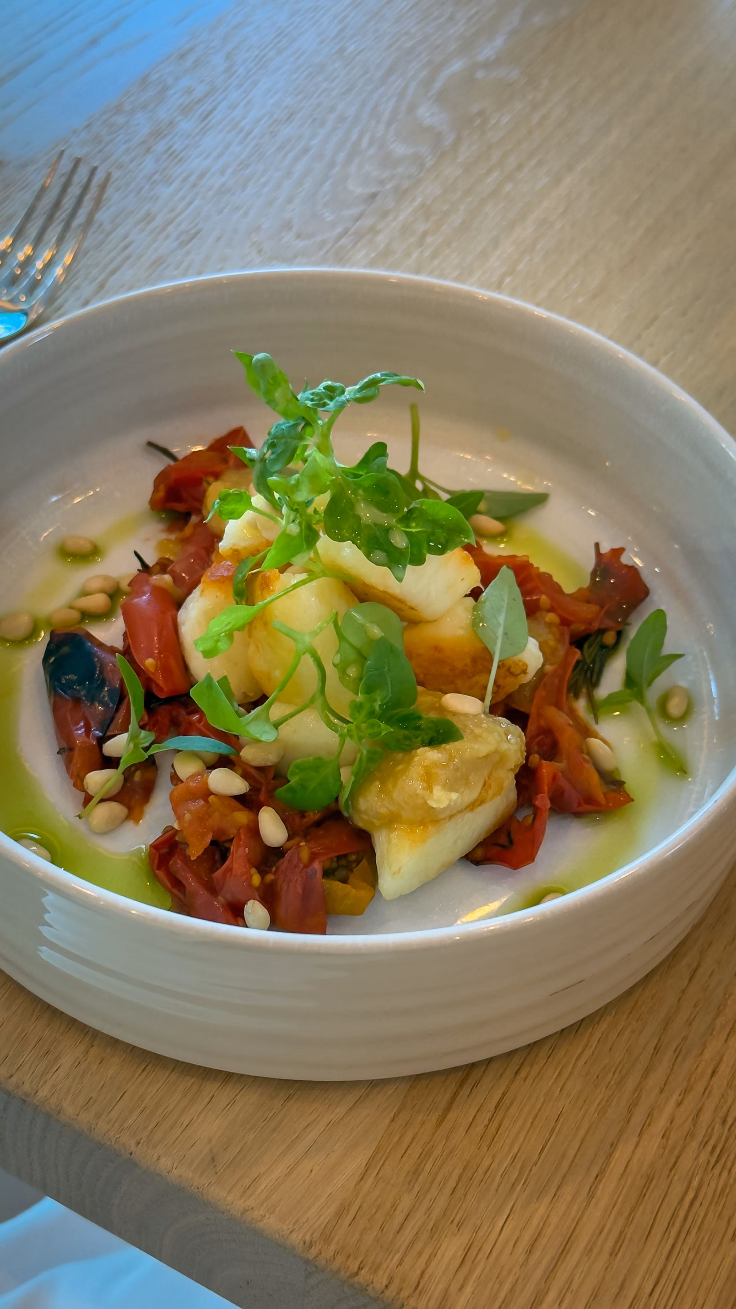 White bowl with a gourmet dish of fried cheese cubes on sautéed red vegetables, topped with green leafy herbs and sprinkled pine nuts, placed on a wooden table.