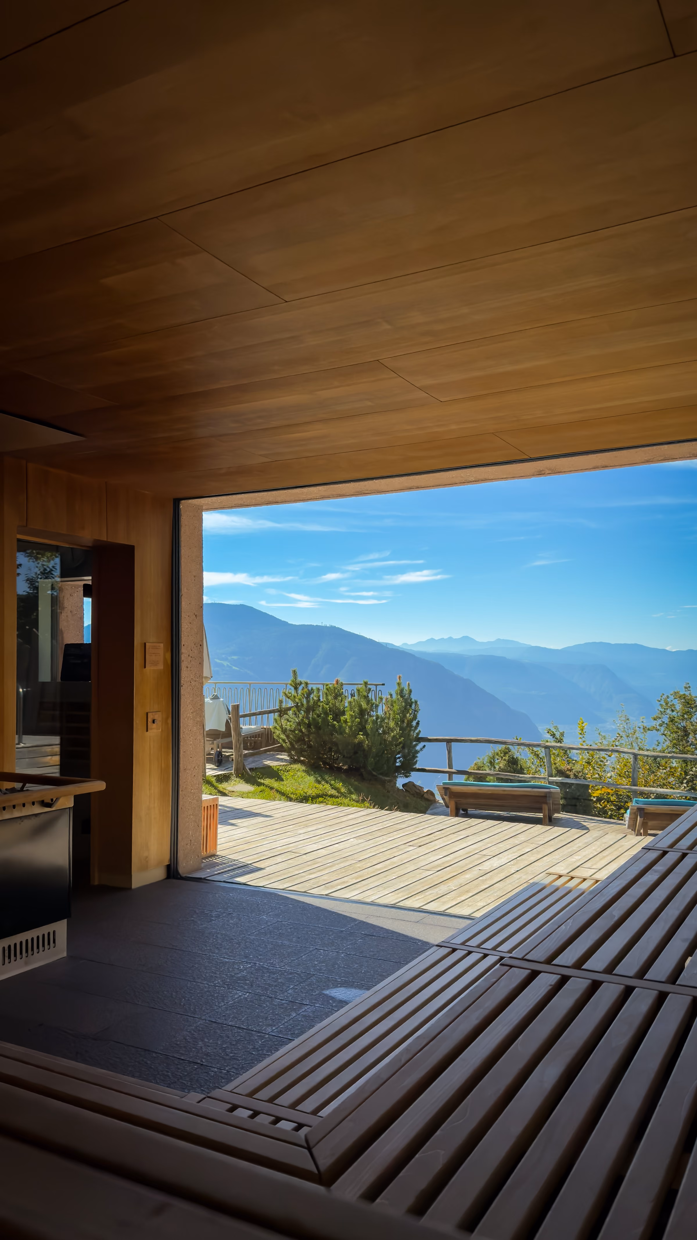 View from inside a wooden cabin looking out through a large open doorway onto a wooden deck with lounge chairs and green bushes, overlooking distant blue mountains under a clear sky.