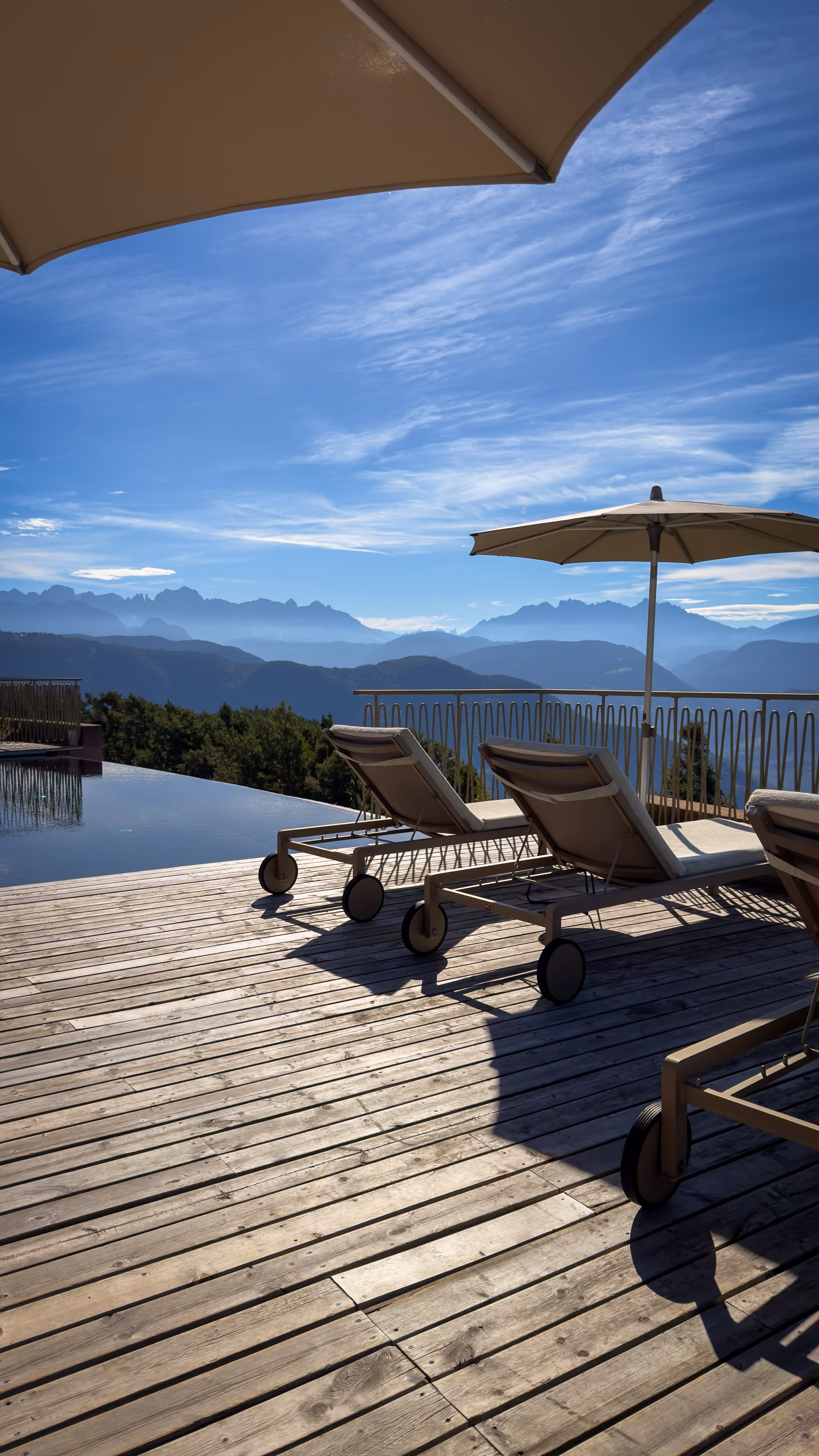 Outdoor wooden deck with lounge chairs and large umbrellas overlooking a mountain range under a clear blue sky with scattered clouds.