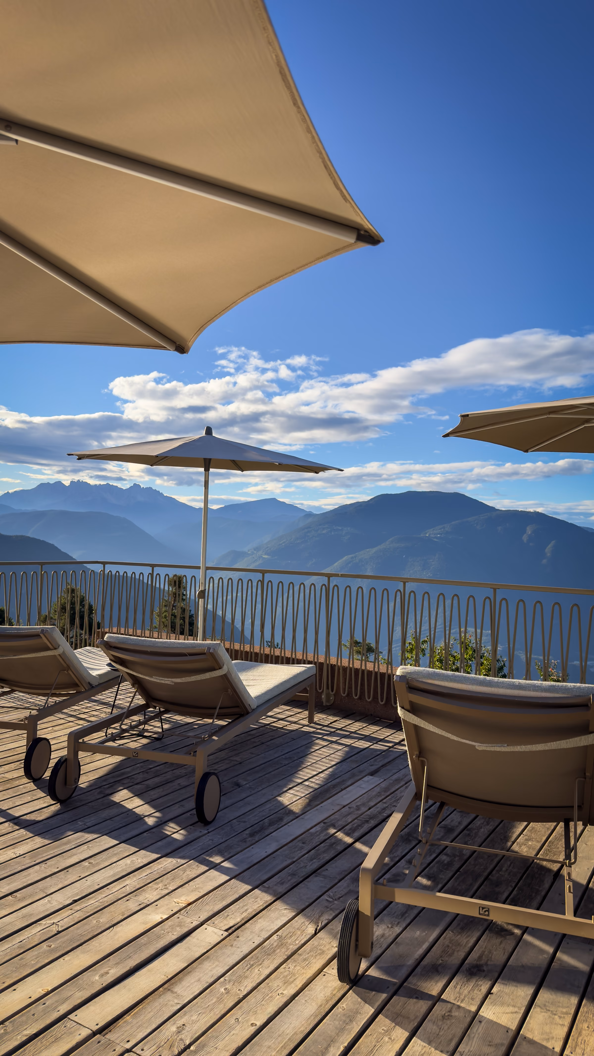 Wooden deck with sun loungers and umbrellas beside an infinity pool overlooking distant mountain ranges under a blue sky.