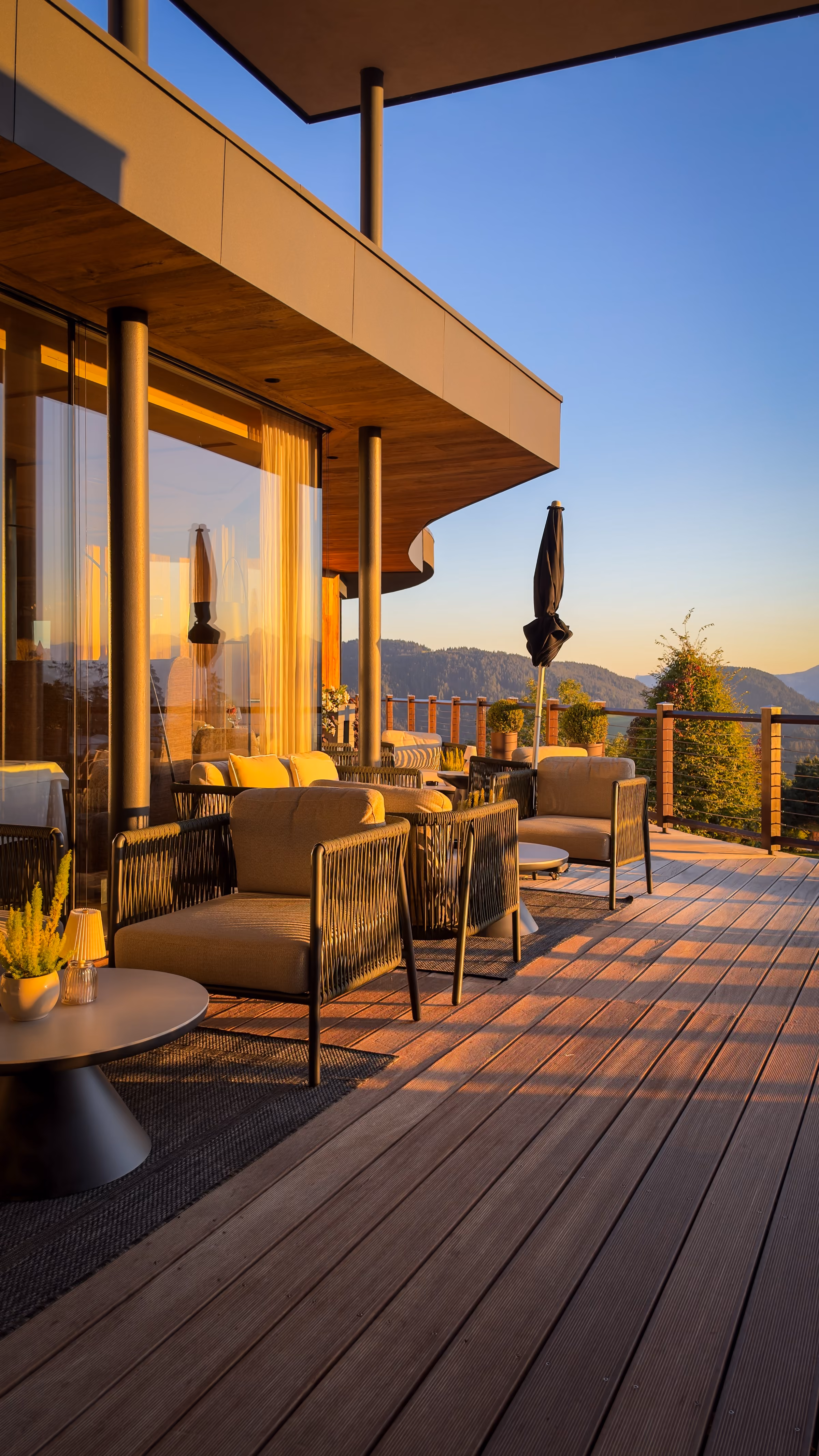 Modern outdoor patio area with wooden decking next to green grass, stone steps covered in ivy, and a large glass window reflecting trees and sunlight.