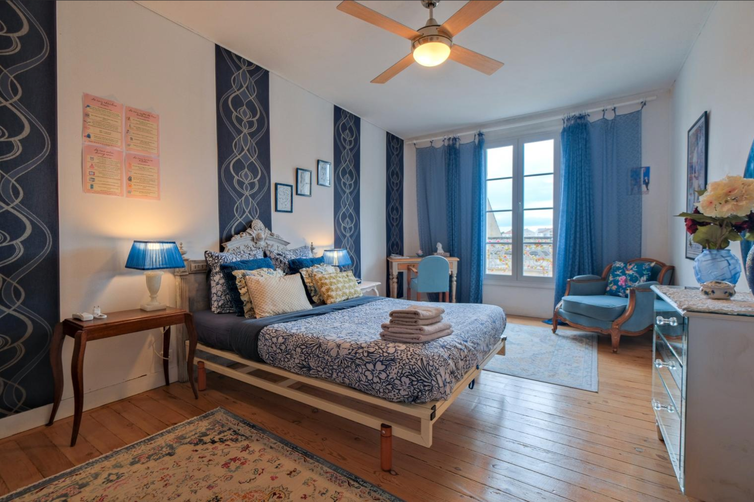 Chambre élégante avec lit double à motif floral bleu et blanc, deux lampes avec abat-jour bleu, bureau avec chaise bleue, fauteuil vintage bleu et grandes fenêtres avec rideaux bleus.
