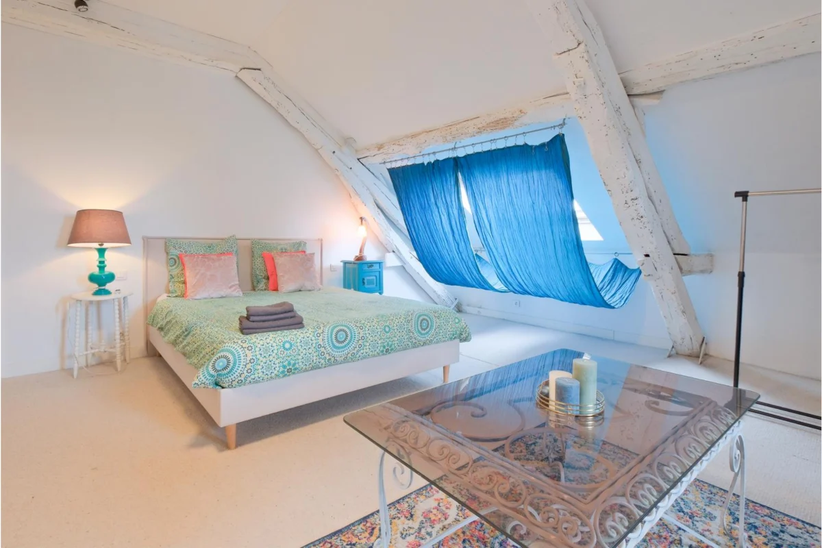 Chambre mansardée avec lit double à couette bleue à motifs, lampes de chevet, rideaux bleus suspendus, et table en verre avec bougies.
