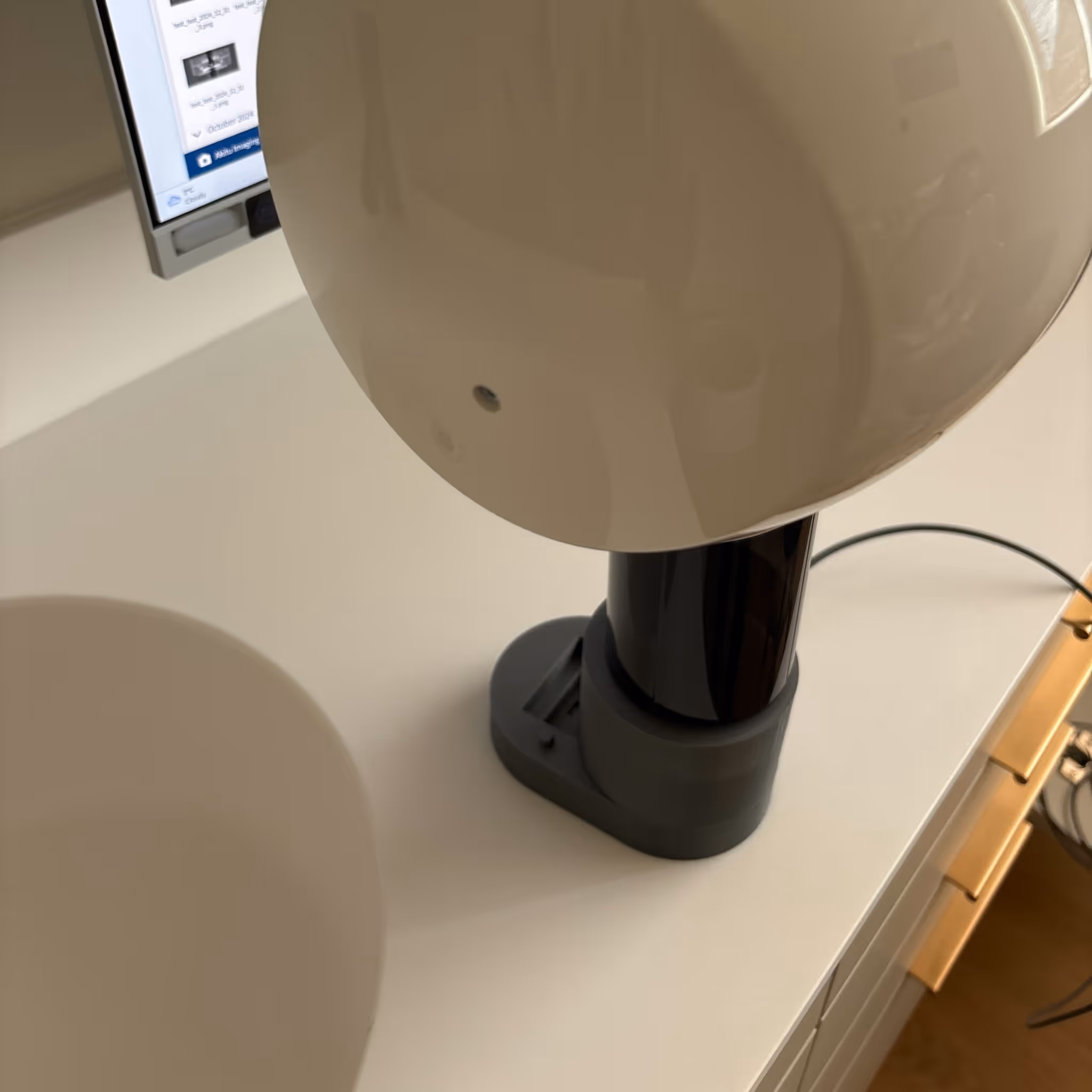 Close-up of a white table lamp with a black base on a white desk with a computer monitor in the background.