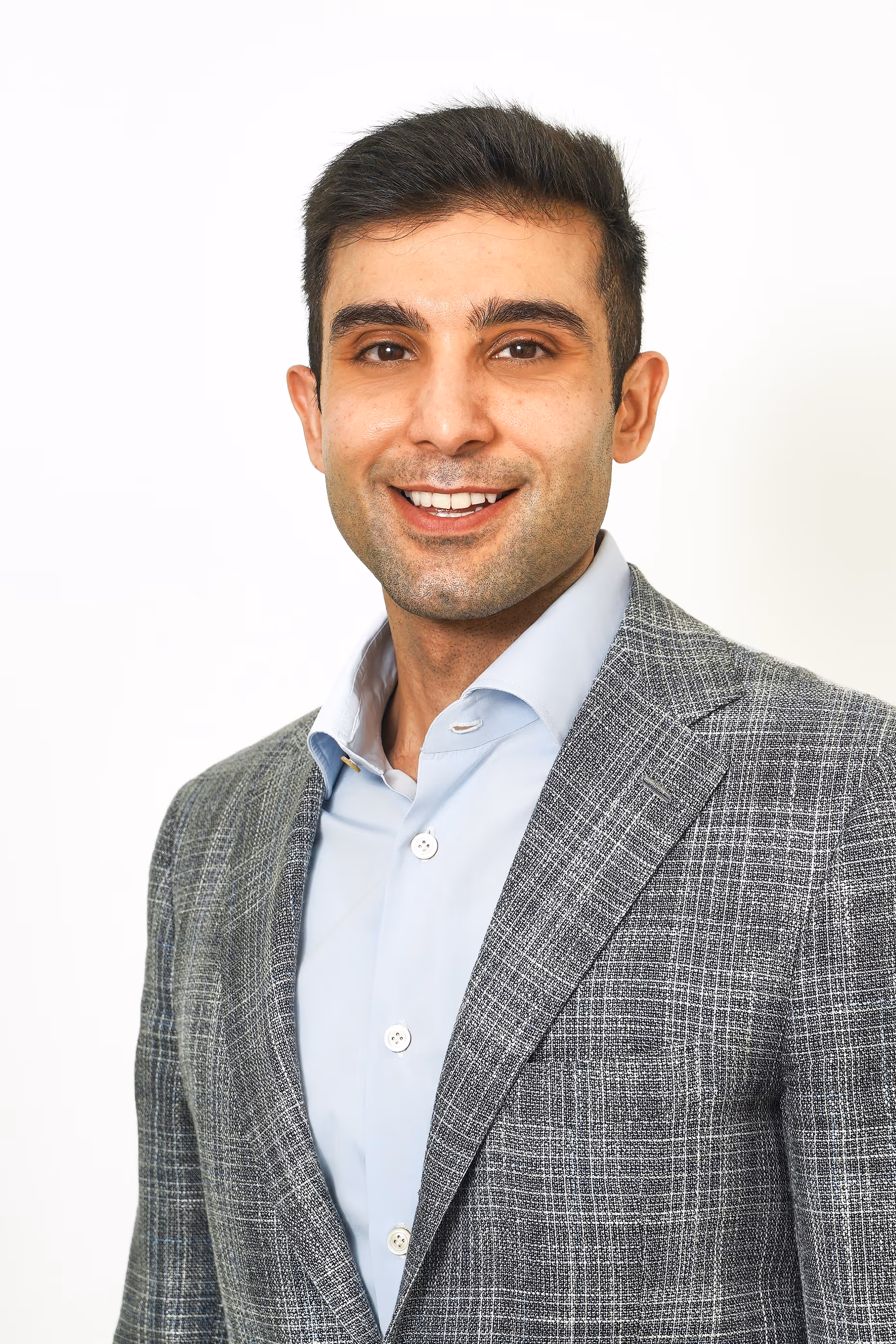 Smiling man wearing a blue shirt and gray checkered blazer against a white background. Fahad Jilani.