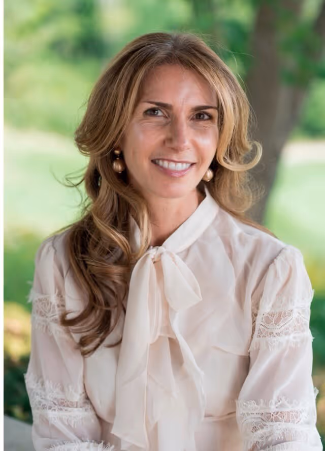 Dr. Nancy Di Santo who has wavy light brown hair and is wearing pearl earrings and a cream blouse with lace and a bow, outdoors.
