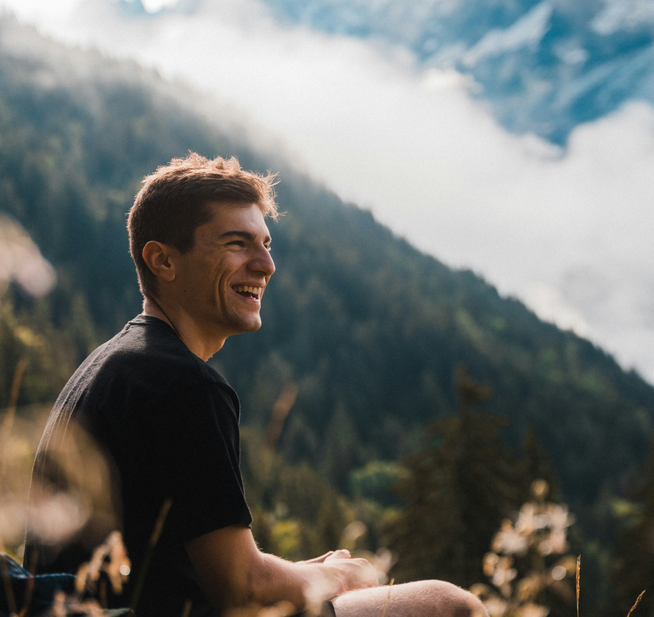 Young man sitting outdoors smiling with forested mountains and cloudy sky in the background.