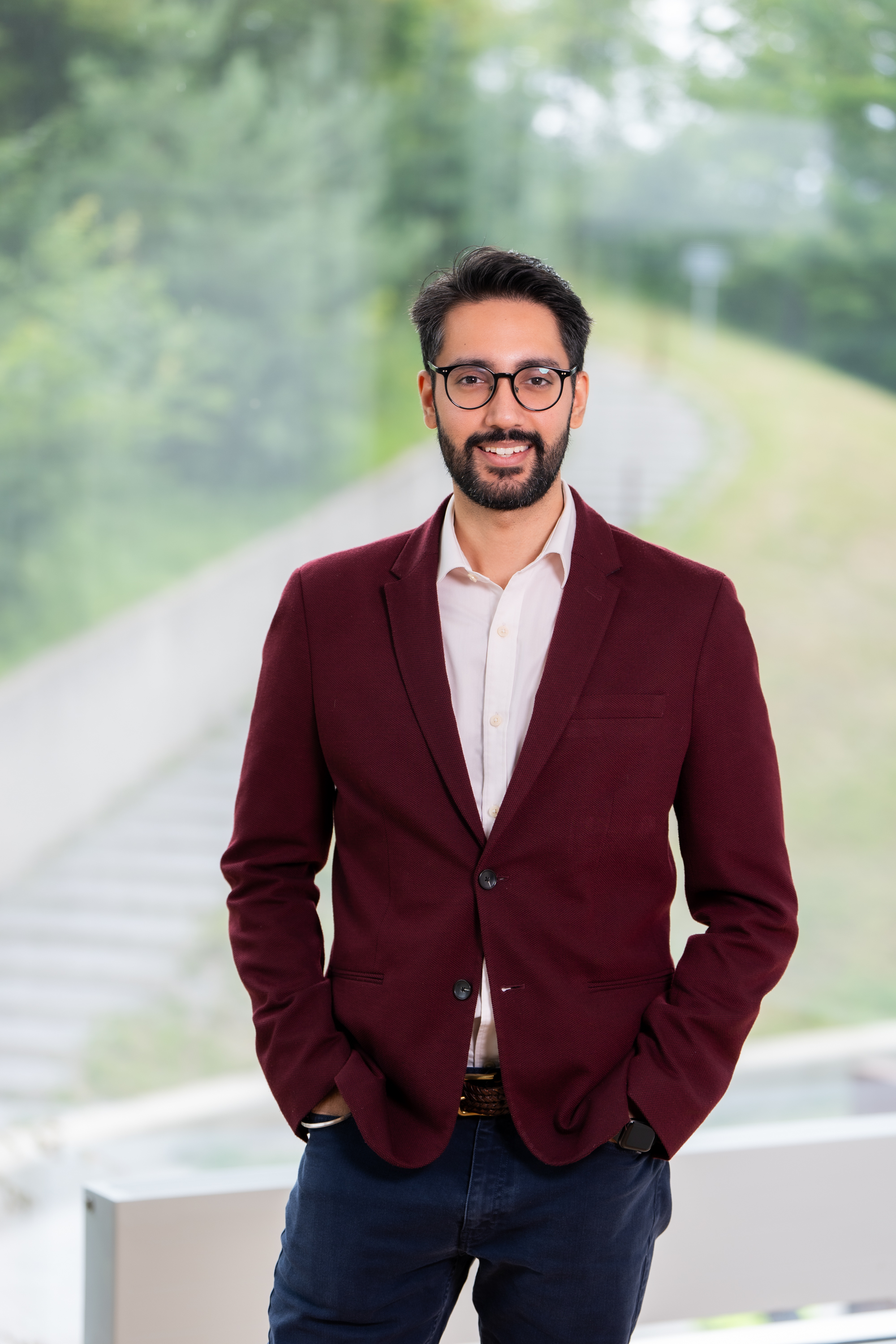 Smiling man with glasses wearing a maroon blazer and white shirt, outdoors with blurred greenery background.
