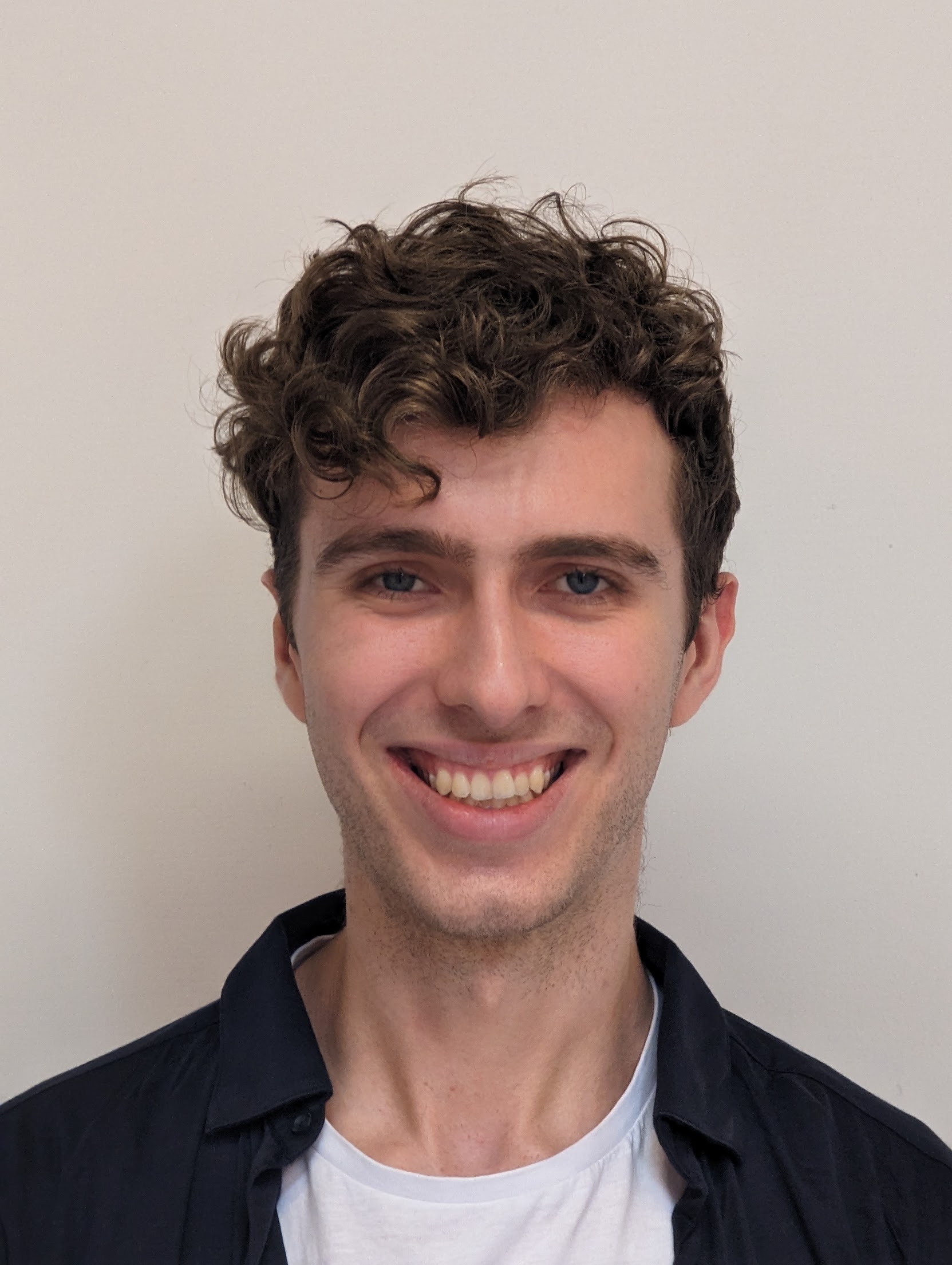 Smiling young man with curly brown hair, blue eyes, wearing a white shirt and dark jacket.