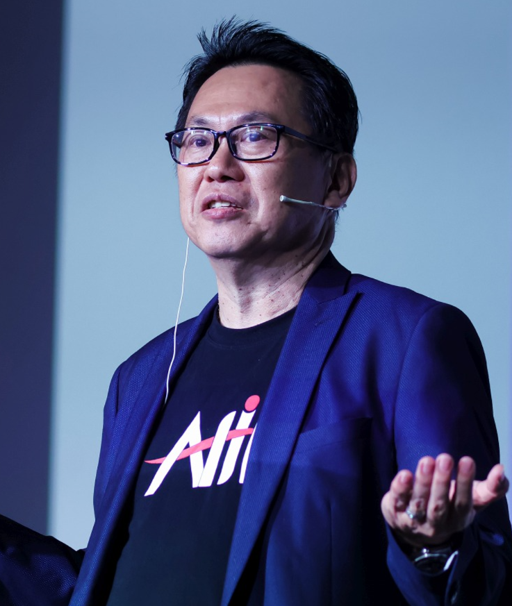 Man wearing glasses, blue blazer, black Ali t-shirt, speaking with microphone headset and hand extended.