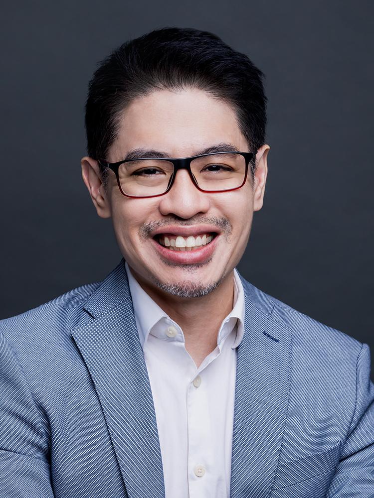 Smiling man wearing glasses, a white shirt, and a blue blazer against a dark background.
