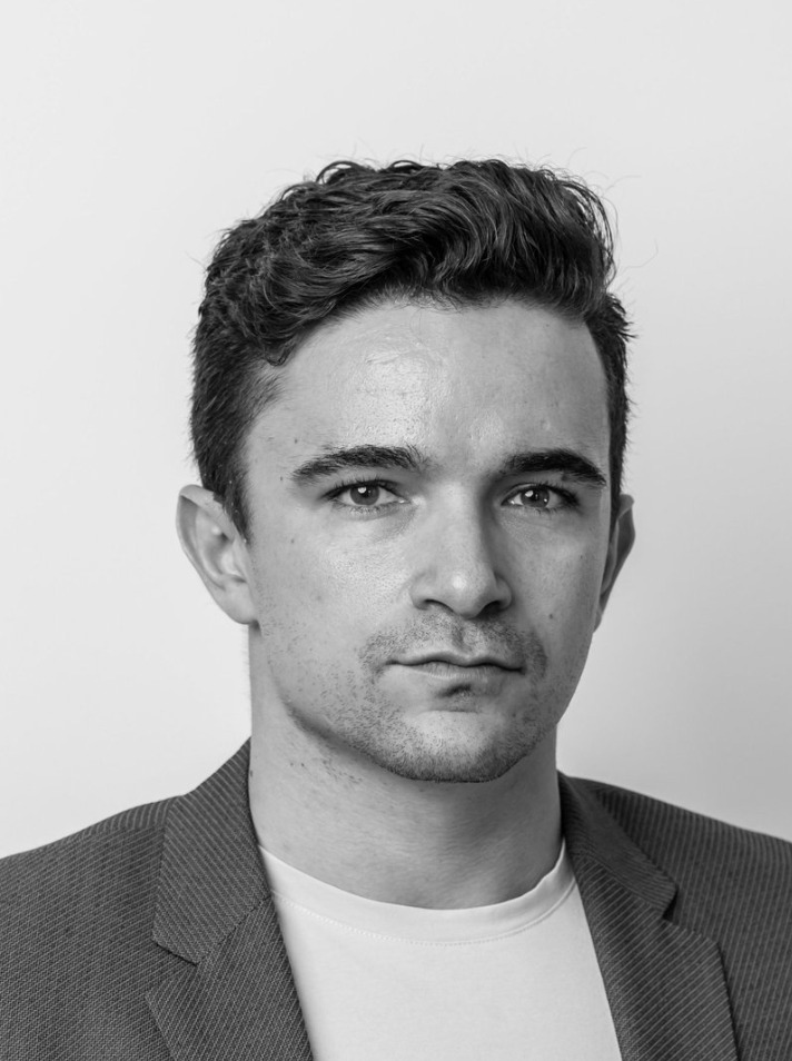 Black and white headshot of a serious young man with short wavy hair wearing a blazer and a t-shirt.
