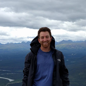 Man in blue shirt and black jacket smiling, standing outdoors with mountains and cloudy sky in background.