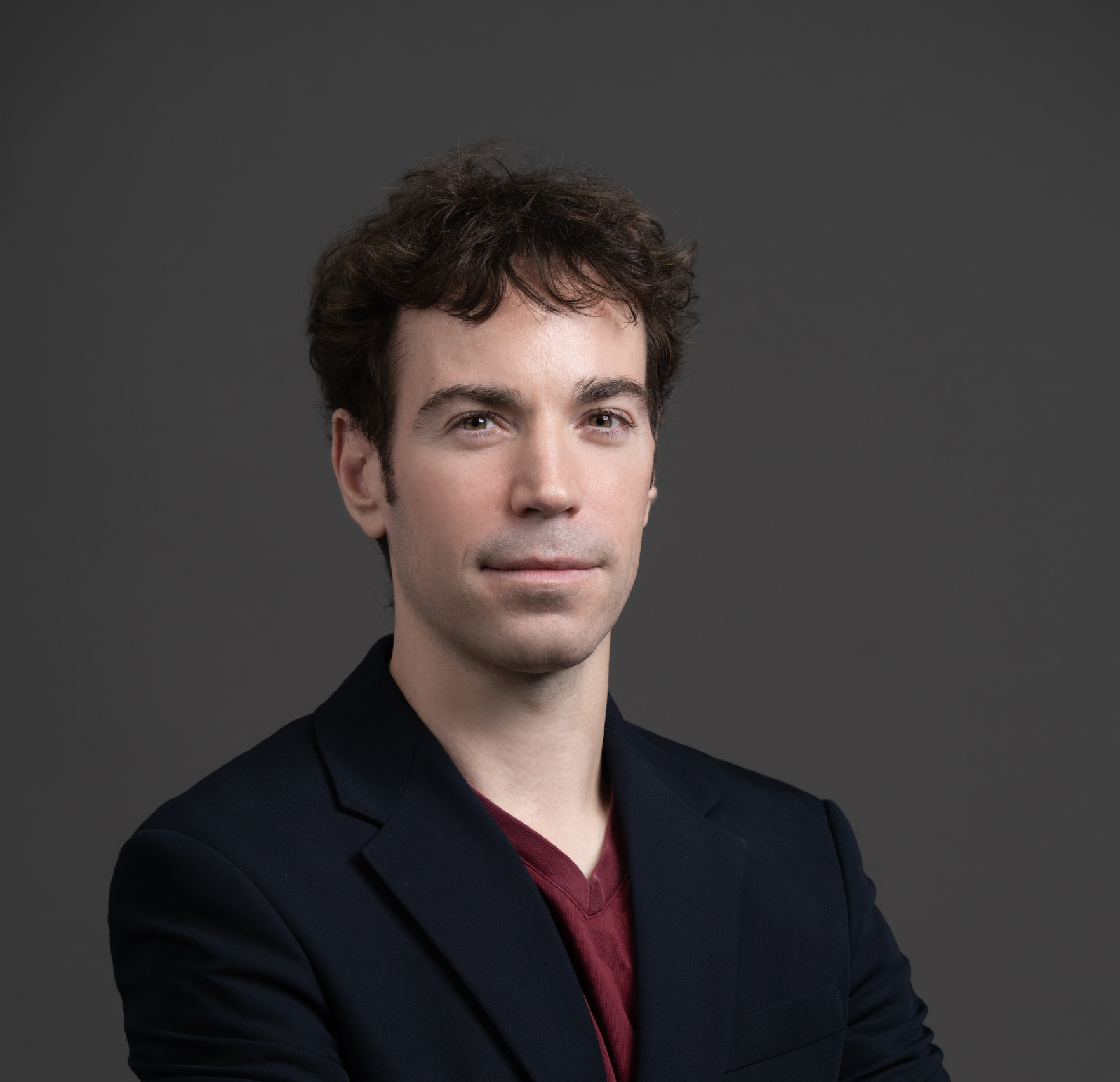Confident young man with curly hair wearing a black blazer and maroon shirt against gray background.
