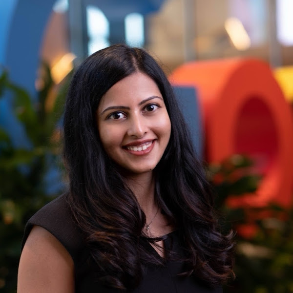 Smiling woman with long dark hair in a black top standing indoors with colorful blurred background.