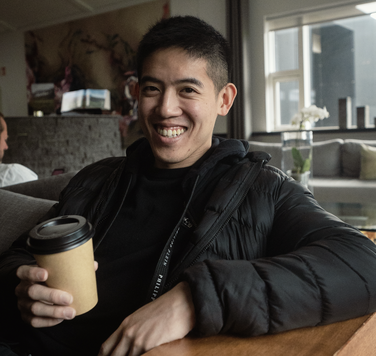 Smiling man in a black jacket holding a takeaway coffee cup while sitting in a cozy room.