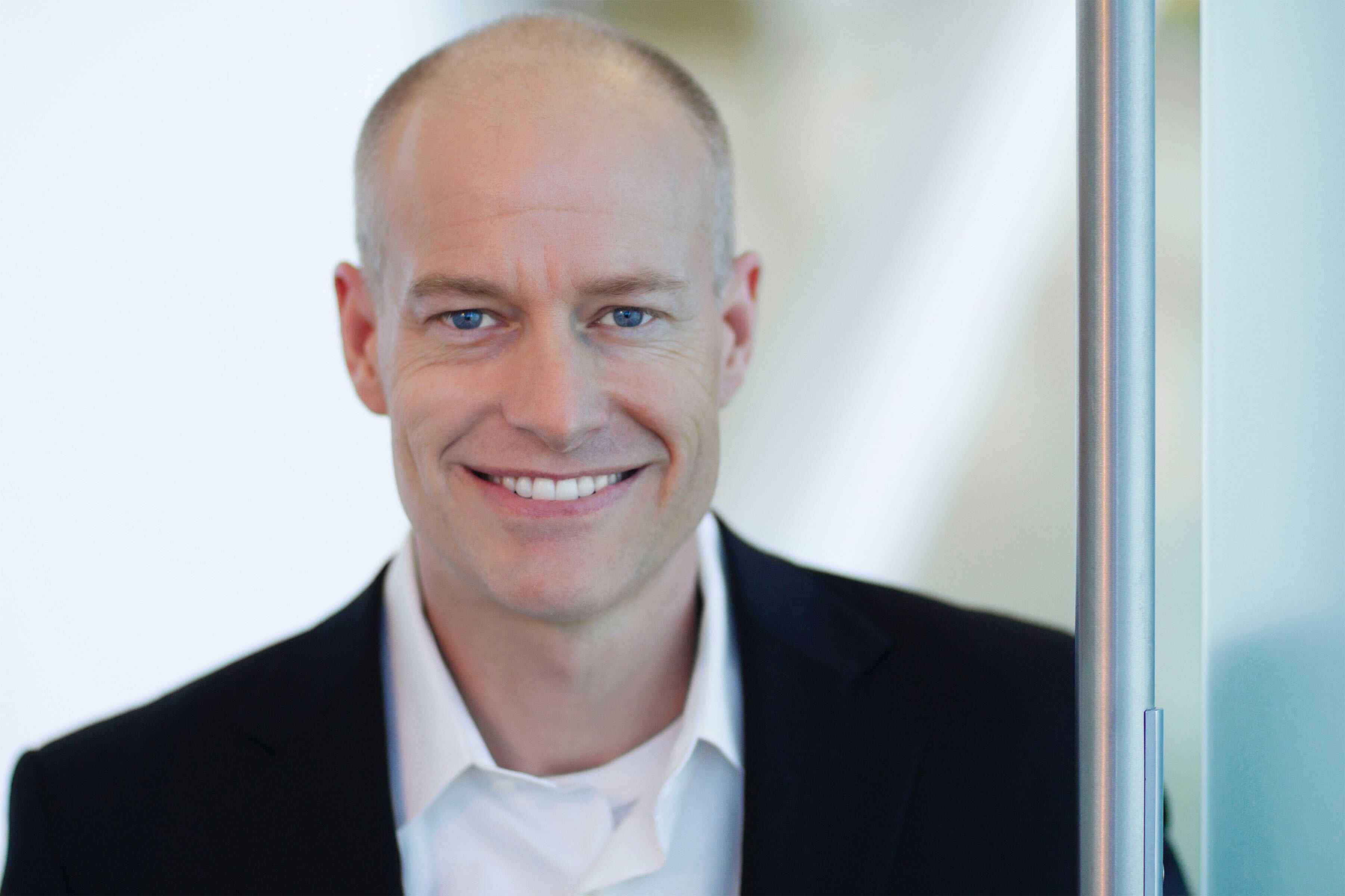Smiling bald man with blue eyes wearing a white shirt and black jacket near a glass door.