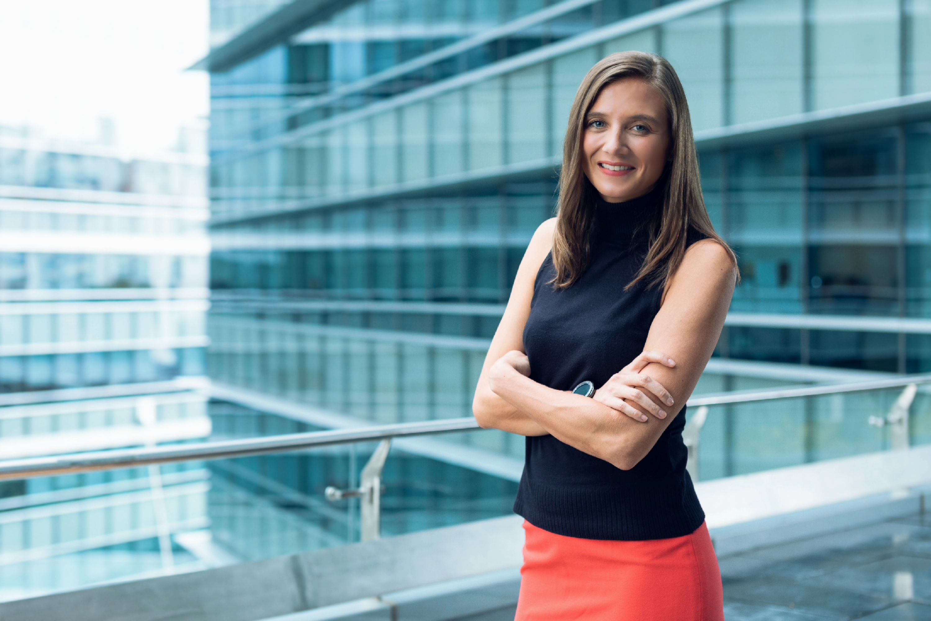 Smiling woman with arms crossed wearing black sleeveless top and red skirt by modern glass building.