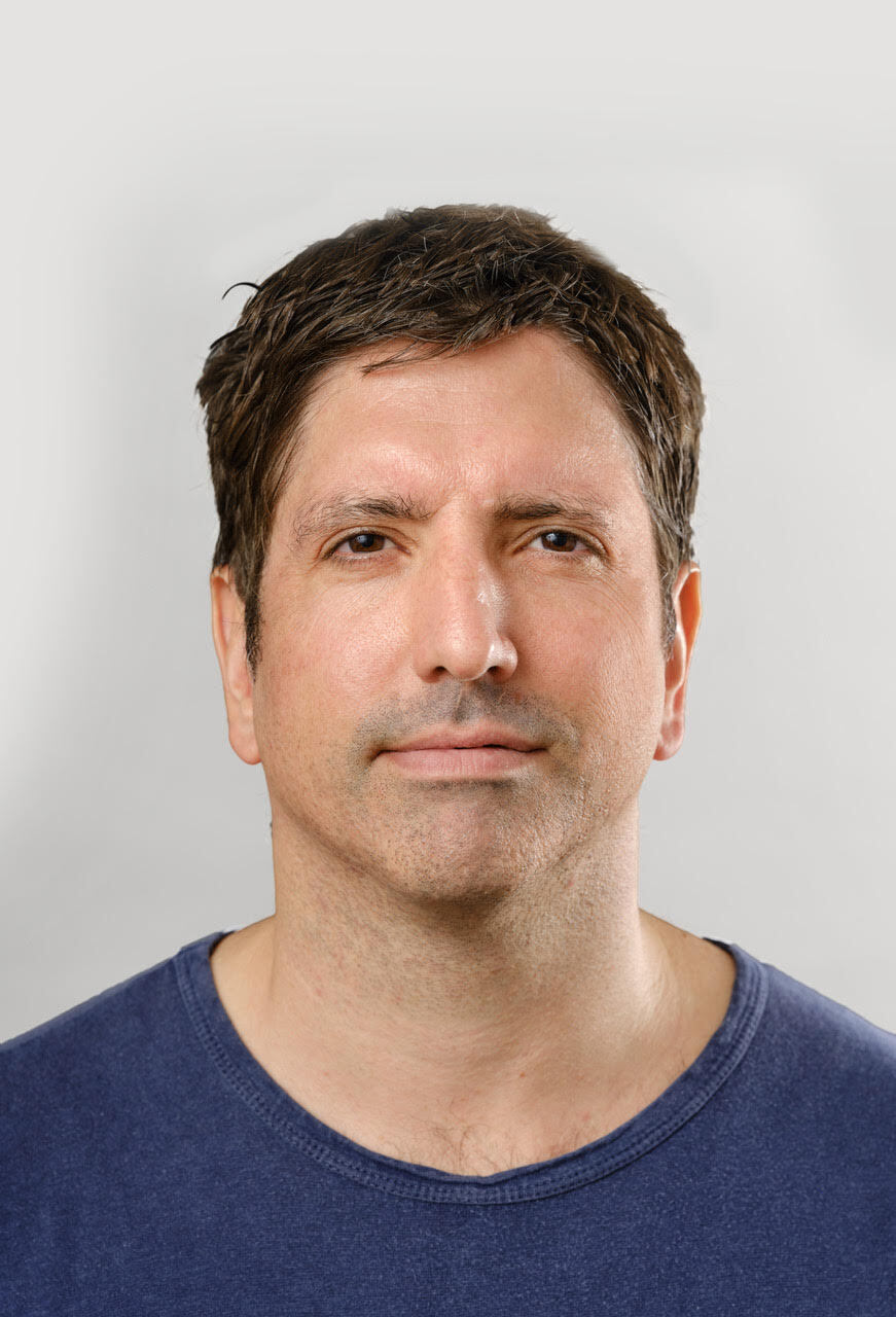 Close-up of a man with short dark hair wearing a navy blue shirt against a light background.