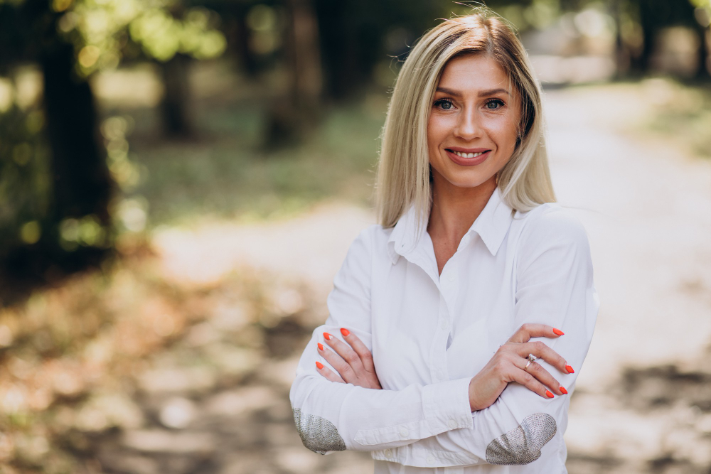 Lächelnde blonde Frau mit weißen Hemd und verschränkten Armen draußen in einem bewaldeten Weg.