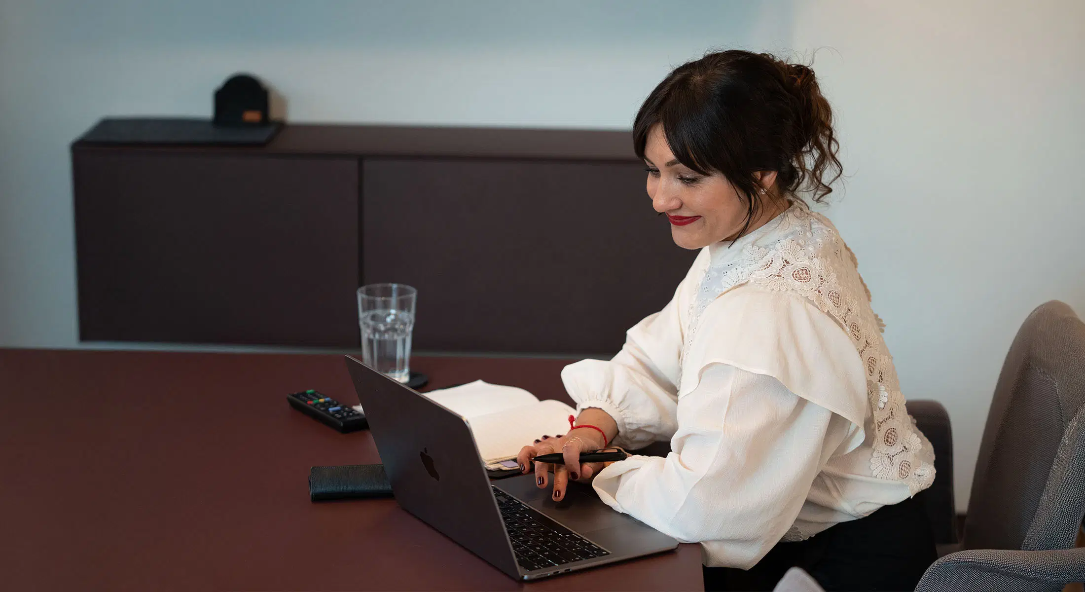 Eine Frau mit einem weißen, bestickten Spitzenoberteil arbeitet an einem Laptop an einem Tisch mit einem Glas Wasser und einem offenen Notizbuch als Unternehmensberatung in München 