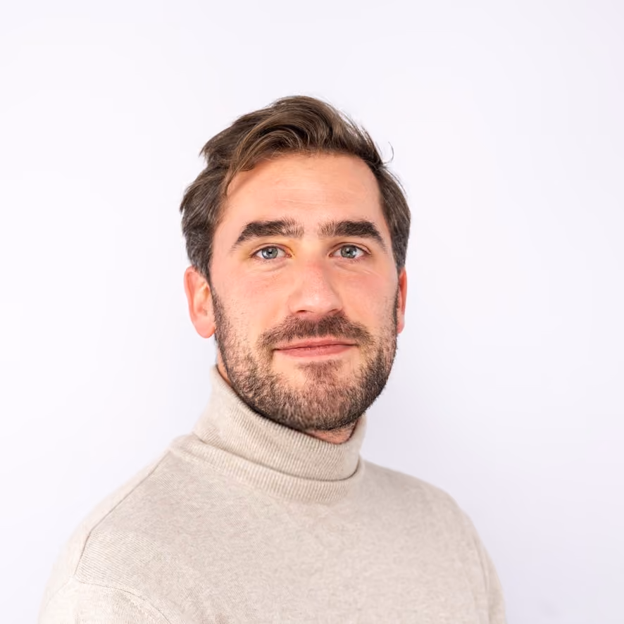 Man with short brown hair and beard wearing a beige turtleneck sweater against a white background.