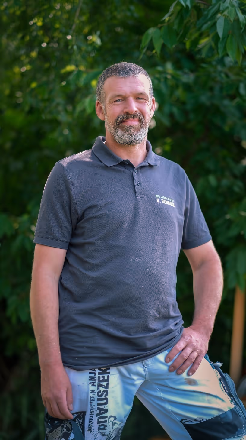 Man with beard in a dark gray polo shirt and paint-stained pants standing outdoors with greenery behind him.